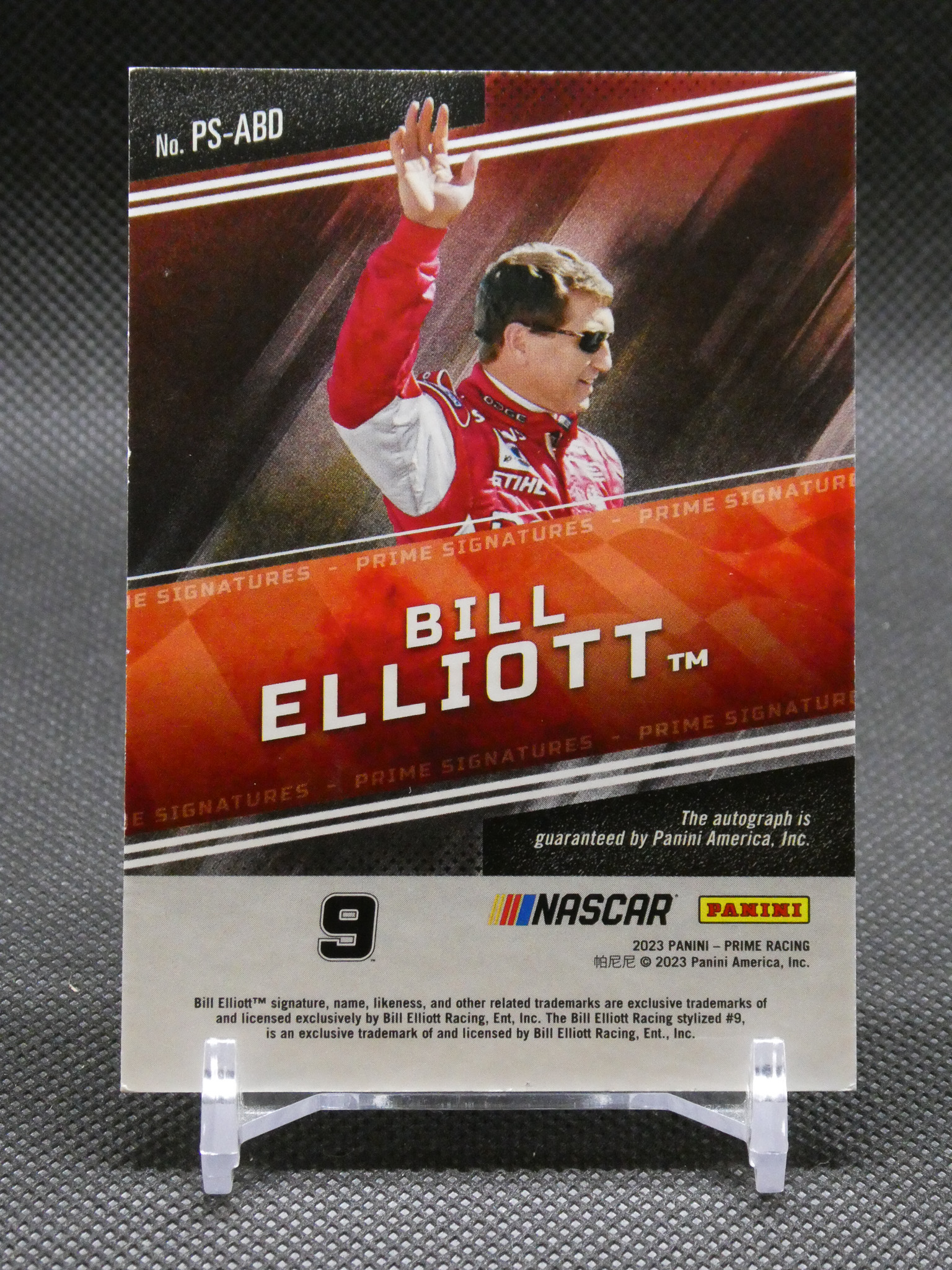 Panini NASCAR trading card of Bill Elliott (#9) with Elliott in a red-and-white racing suit, raising his hand, on a display stand.