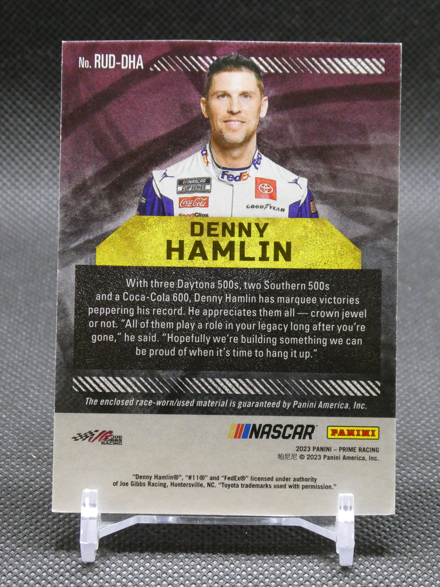 Collectible NASCAR trading card featuring driver Denny Hamlin in a white racing suit with a yellow name banner and race text below.