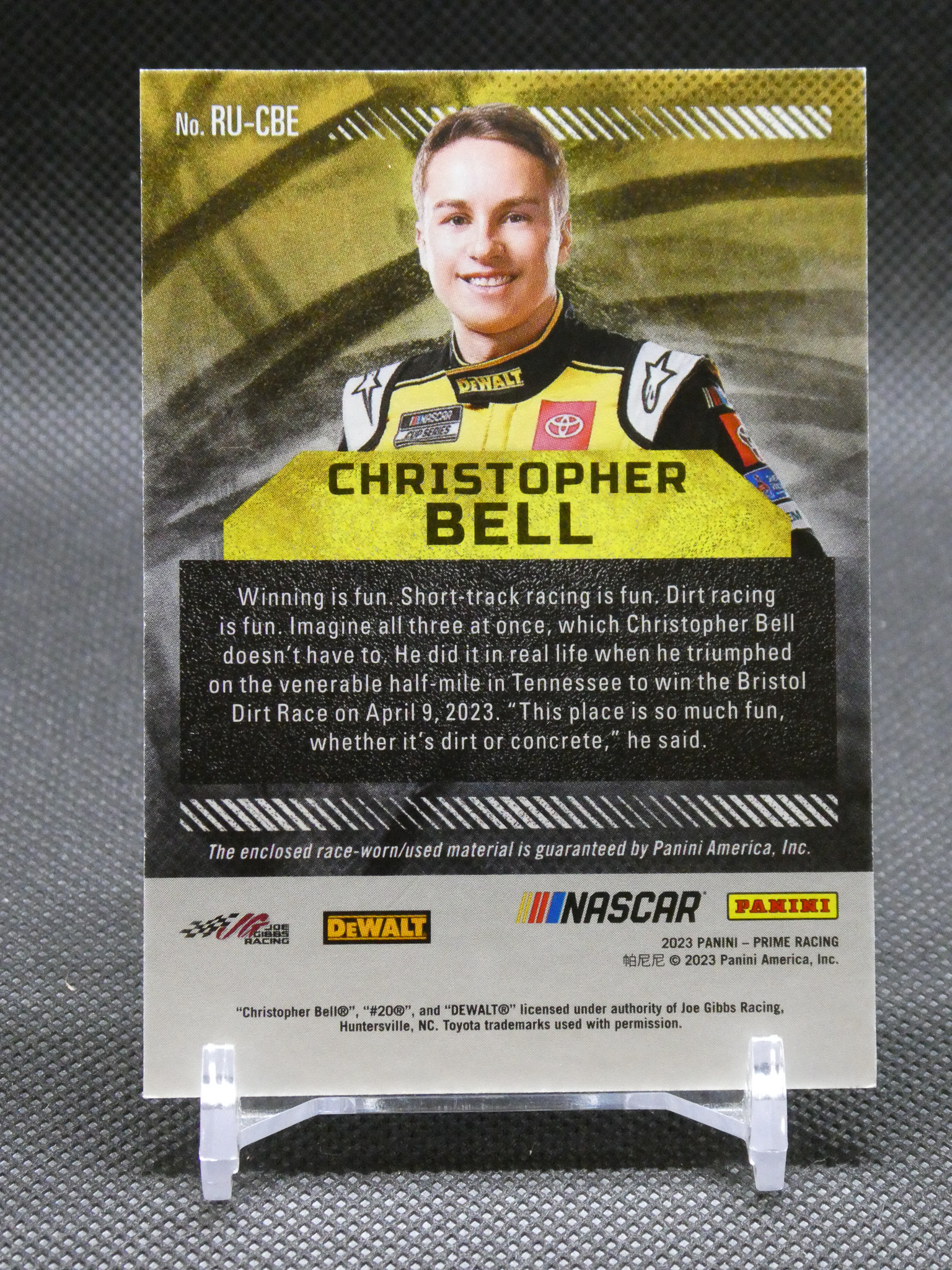 Trading card of NASCAR driver Christopher Bell in a yellow racing suit, his name banner across the middle, with sponsor logos and a racing quote below.