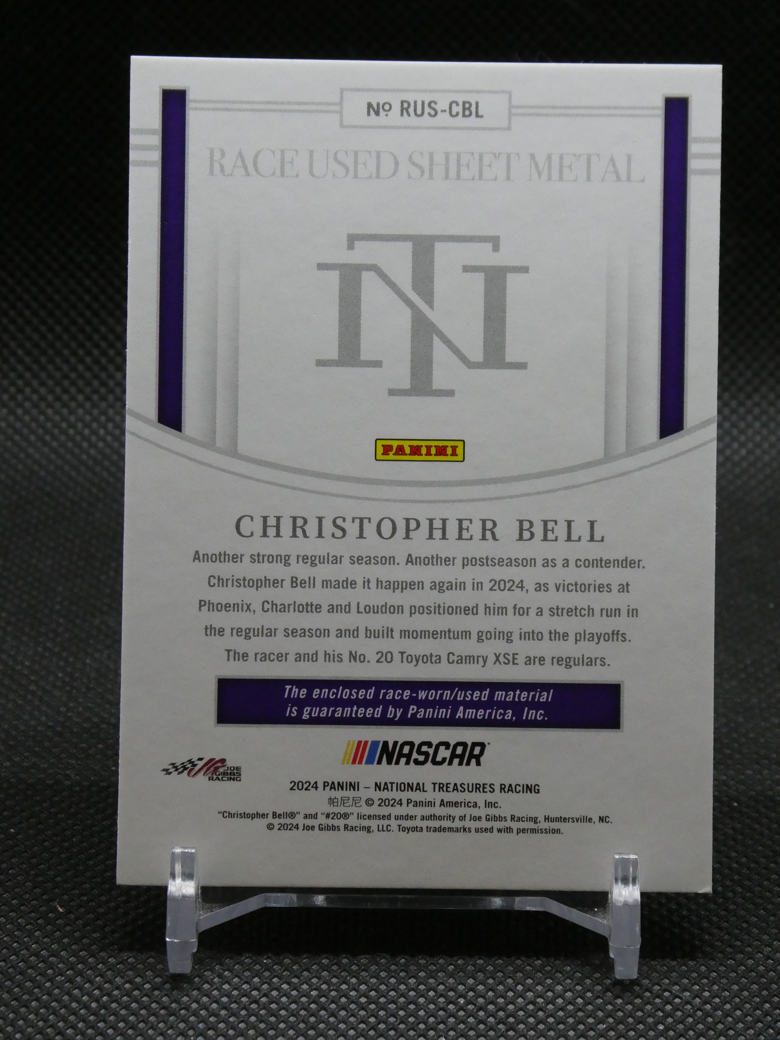Panini NASCAR card honoring Christopher Bell, titled 'Race Used Sheet Metal' with a large interlocking logo and purple accents on a display stand.
