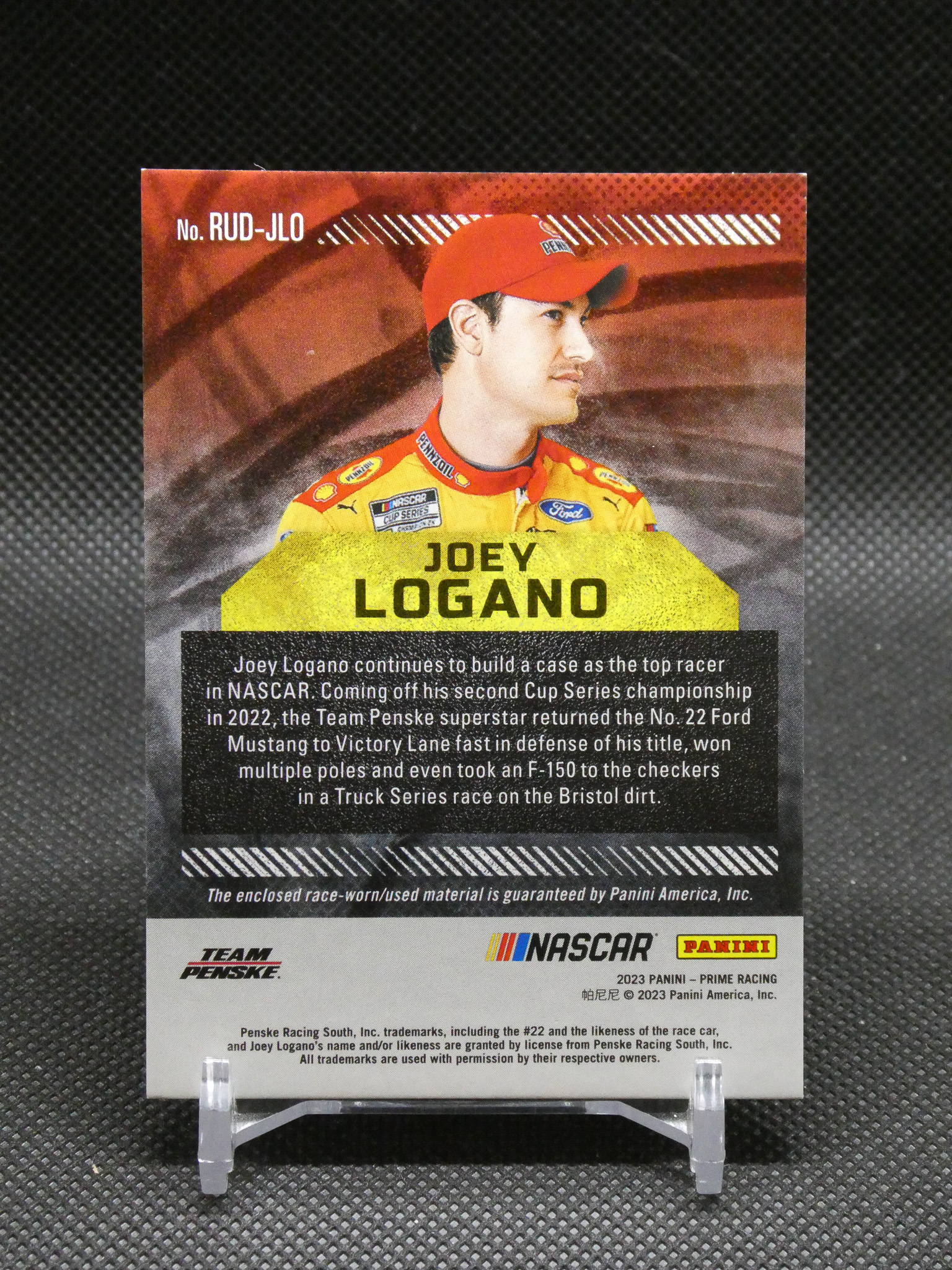 Trading card showing NASCAR driver Joey Logano in a red cap and yellow Penske racing suit with his name on a yellow banner.