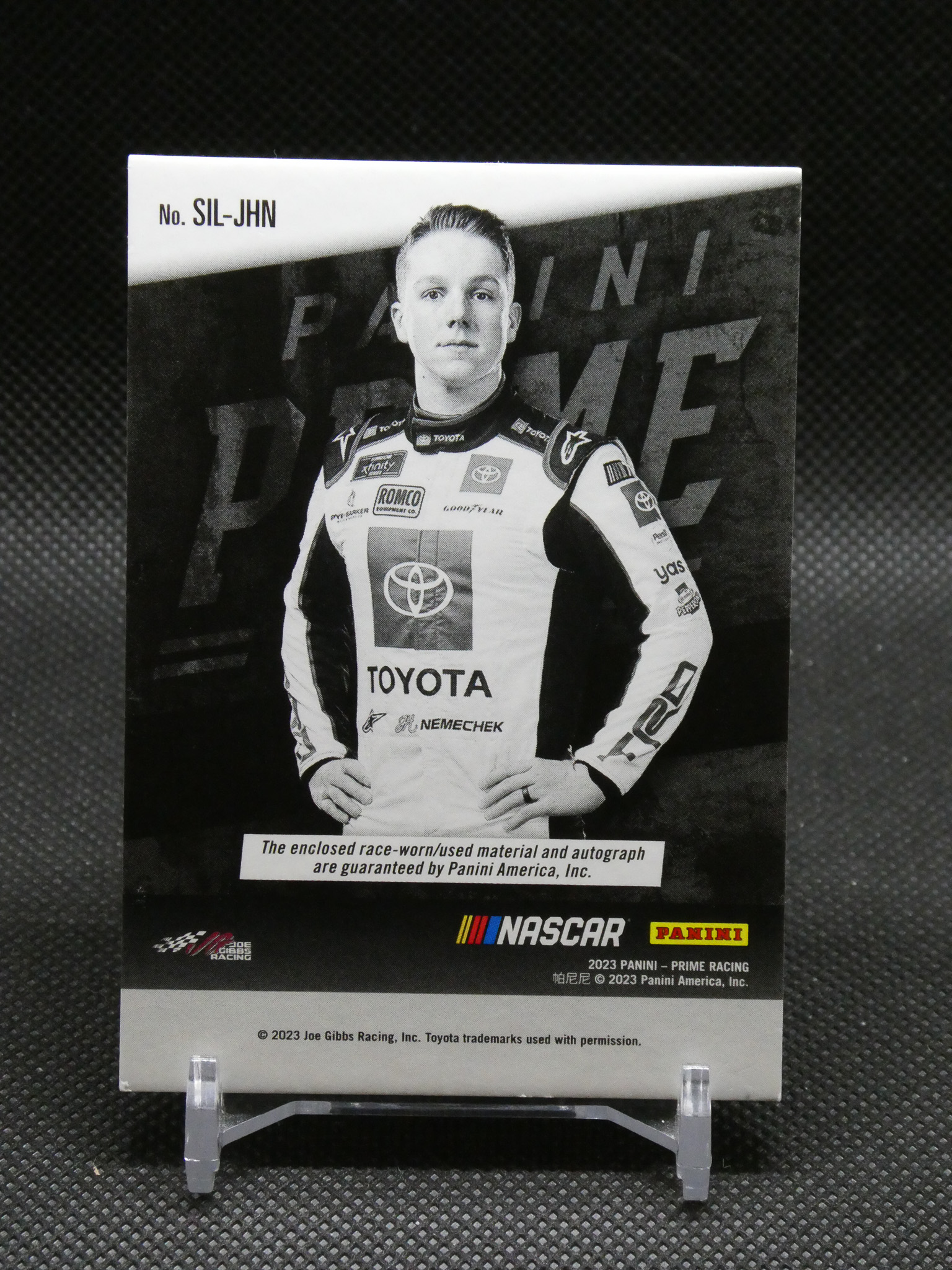Black-and-white NASCAR trading card portrait of a driver in a Toyota race suit, standing with hands on hips, Panini and NASCAR logos visible.