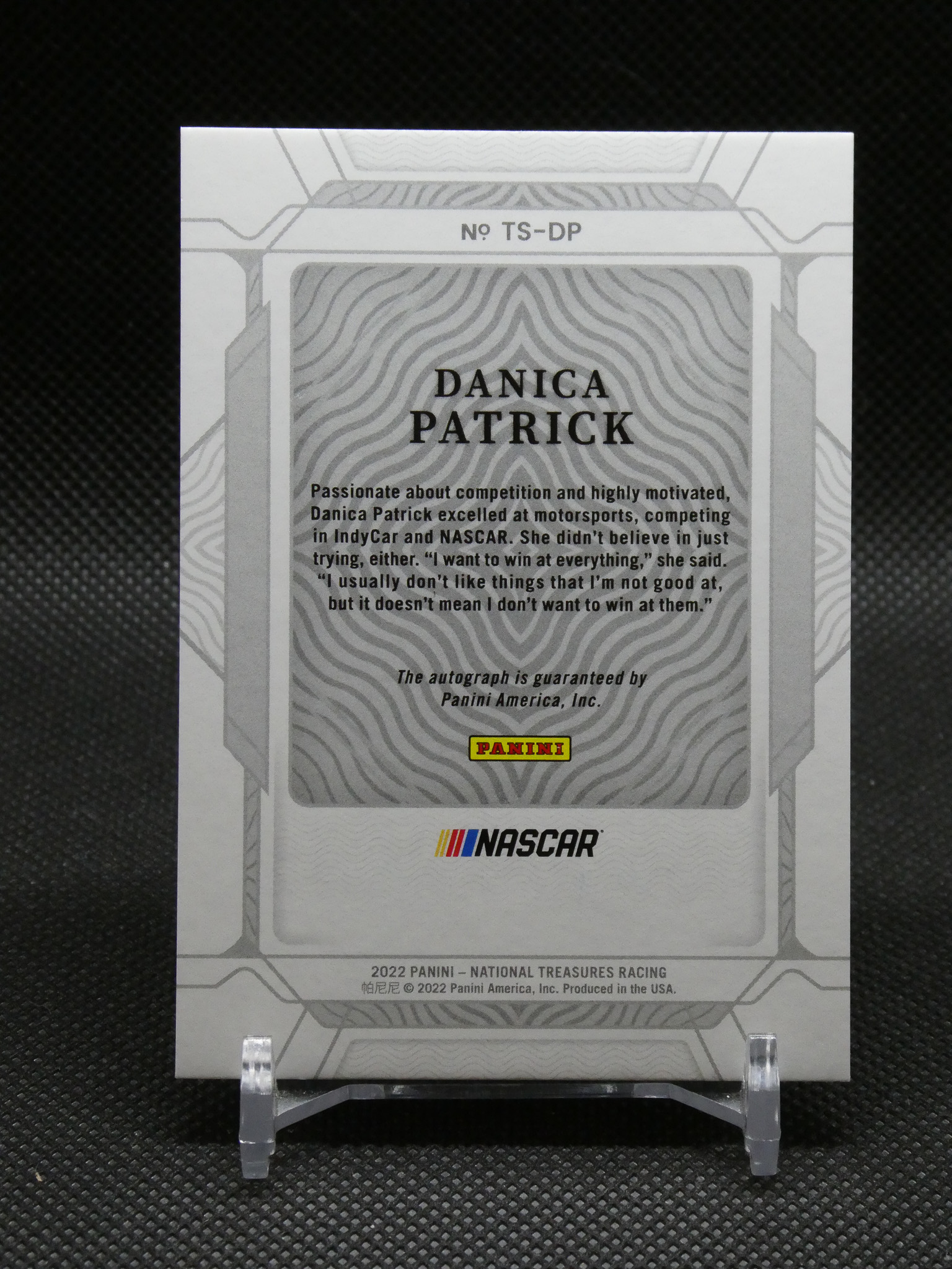 Front of a Panini NASCAR trading card featuring Danica Patrick, with a wavy gray pattern and Panini logo, displayed on a small clear stand on a dark backdrop.