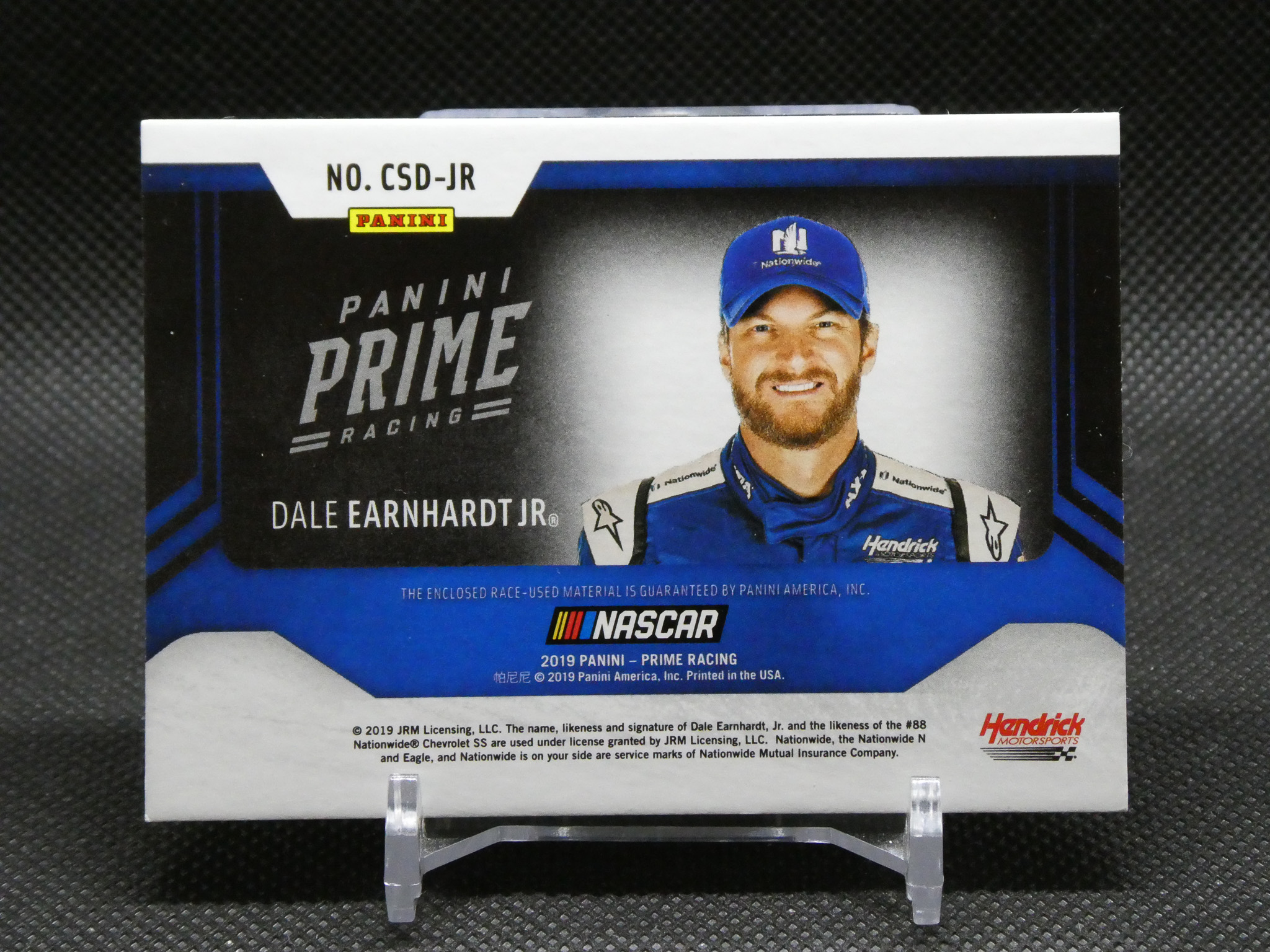 Panini Prime Racing trading card featuring Dale Earnhardt Jr. in a blue cap and racing suit, smiling portrait.