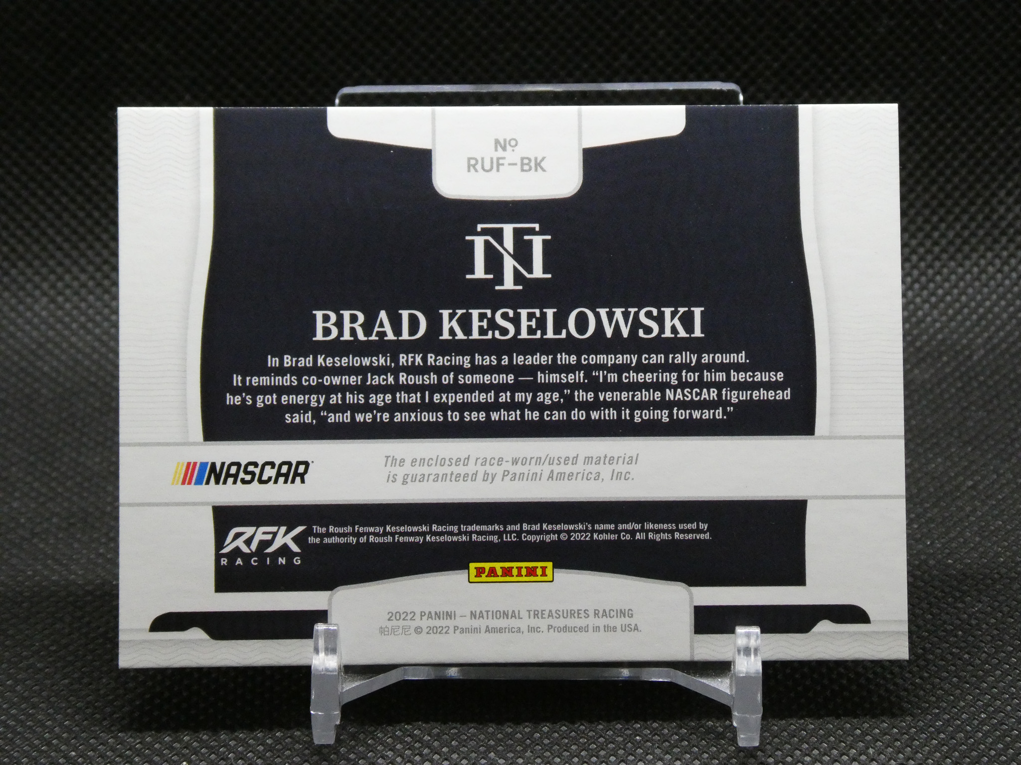 Brad Keselowski NASCAR trading card on a display stand, featuring logos for NASCAR, RFK Racing, and Panini on a dark background.