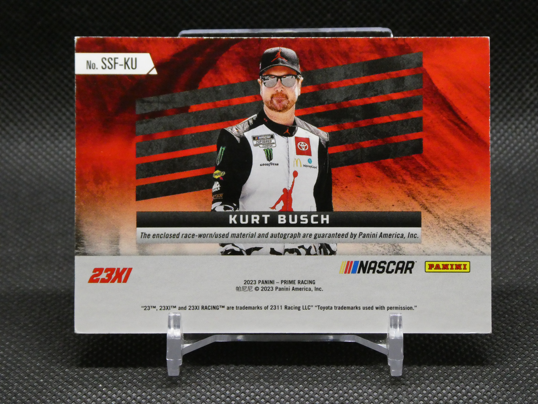 Panini NASCAR trading card featuring Kurt Busch in a black-and-white racing suit with red background and diagonal stripes; shows his name on a black banner and 23XI logo on the left.