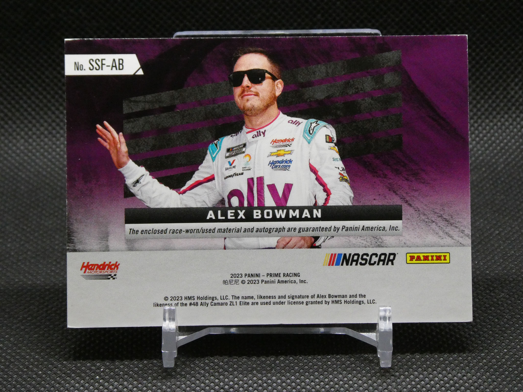 Trading card showing NASCAR driver Alex Bowman in a white racing suit and sunglasses, against a purple background, with his name band across the middle and sponsor logos.