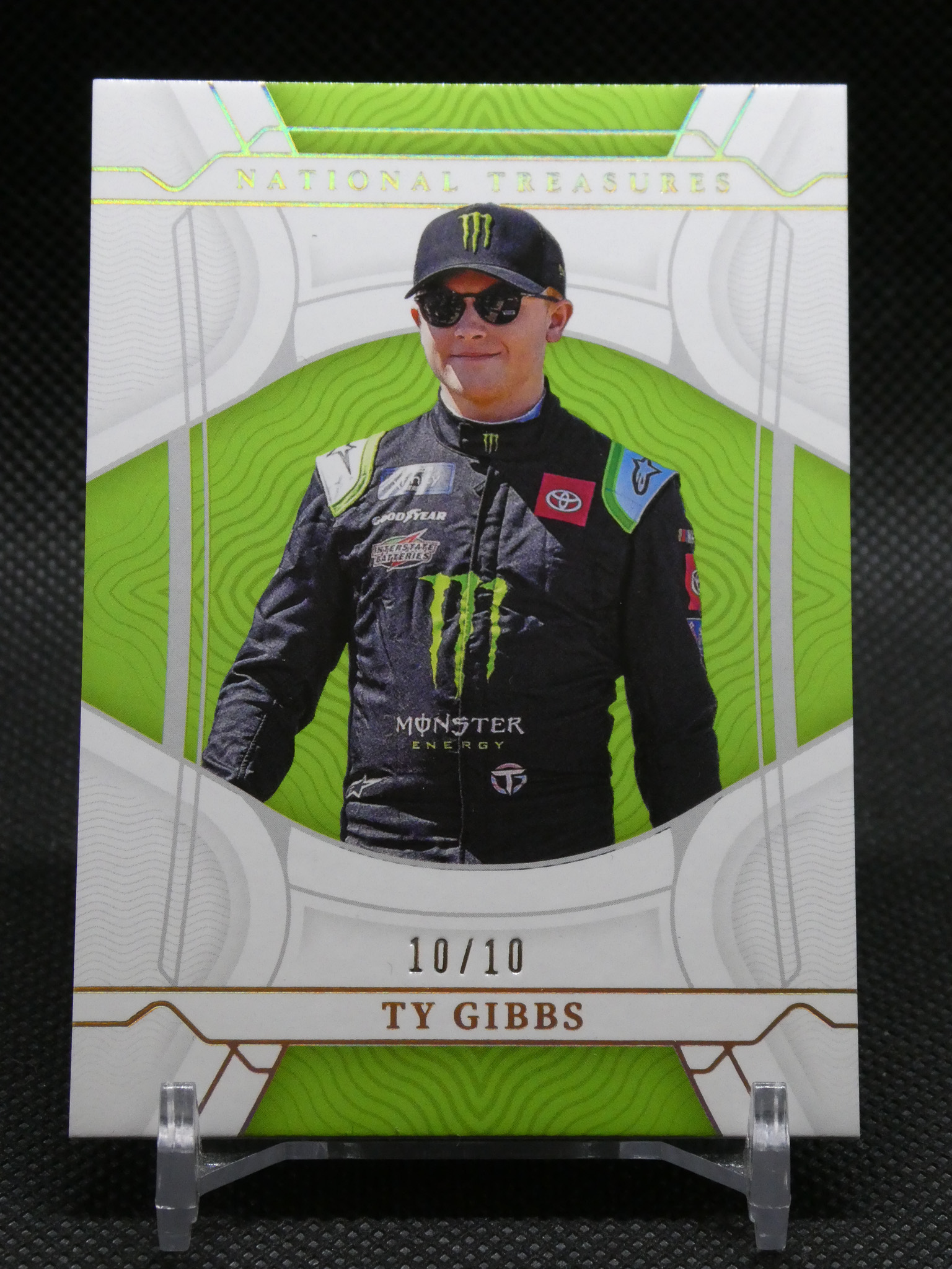 National Treasures trading card of Ty Gibbs, driver in a black Monster Energy suit with green decorative background and sunglasses.