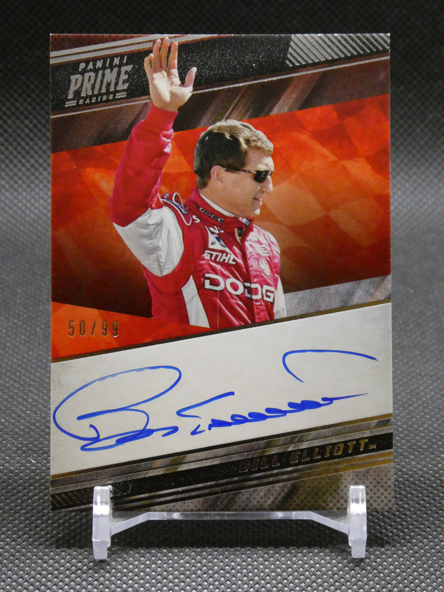Autographed Panini Prime NASCAR card of Bill Elliott, shown in a red racing suit with sunglasses, waving his hand on a red background, on a display stand.