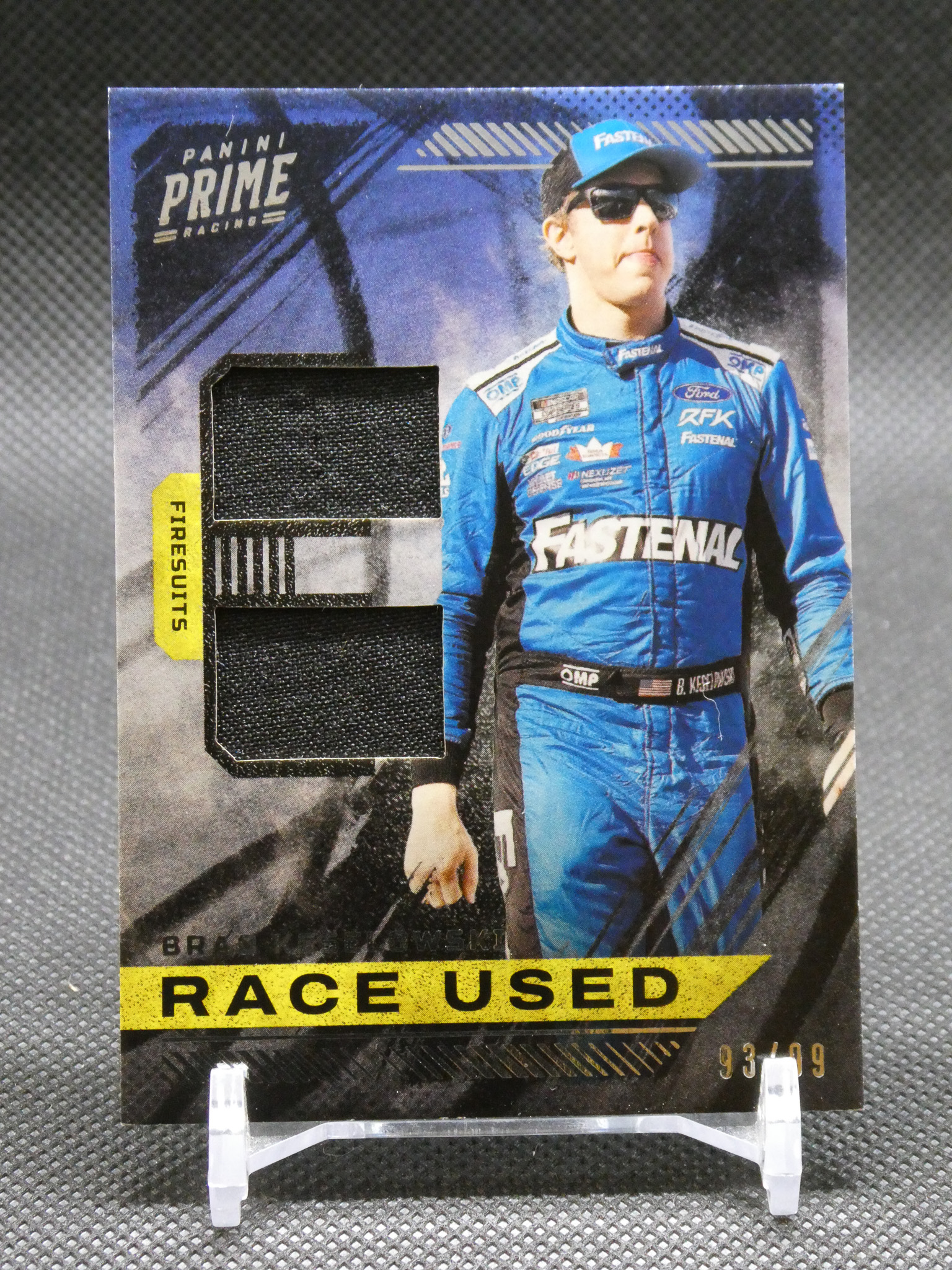 Panini Prime Racing collectible card showing a driver in a blue racing suit with a fabric swatch and a bold 'RACE USED' banner at the bottom.