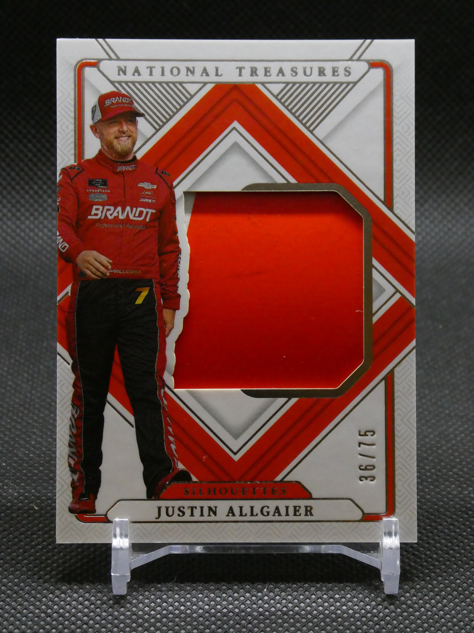 Trading card featuring NASCAR driver Justin Allgaier in a red racing suit with a red geometric cutout window and National Treasures branding above the card edge.