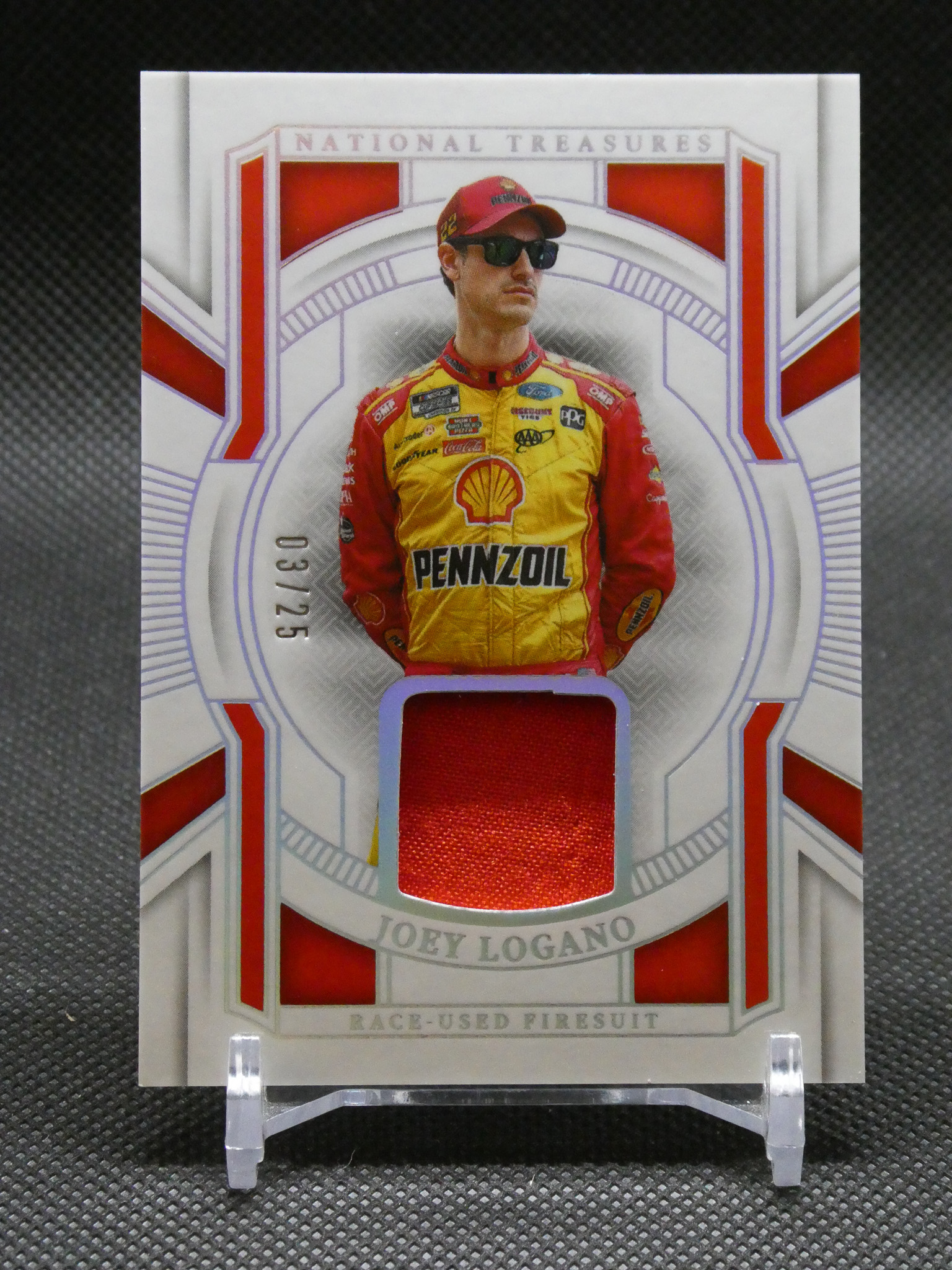 Trading card featuring NASCAR driver Joey Logano in a yellow Pennzoil fire suit with red sleeves, wearing sunglasses and cap, on a decorative background.