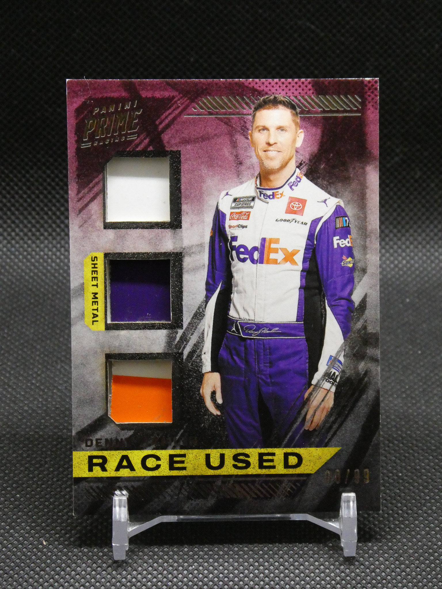 Racing memorabilia card showing a driver in a purple-white suit with race-used fabric swatches on the left and 'RACE USED' text at the bottom
