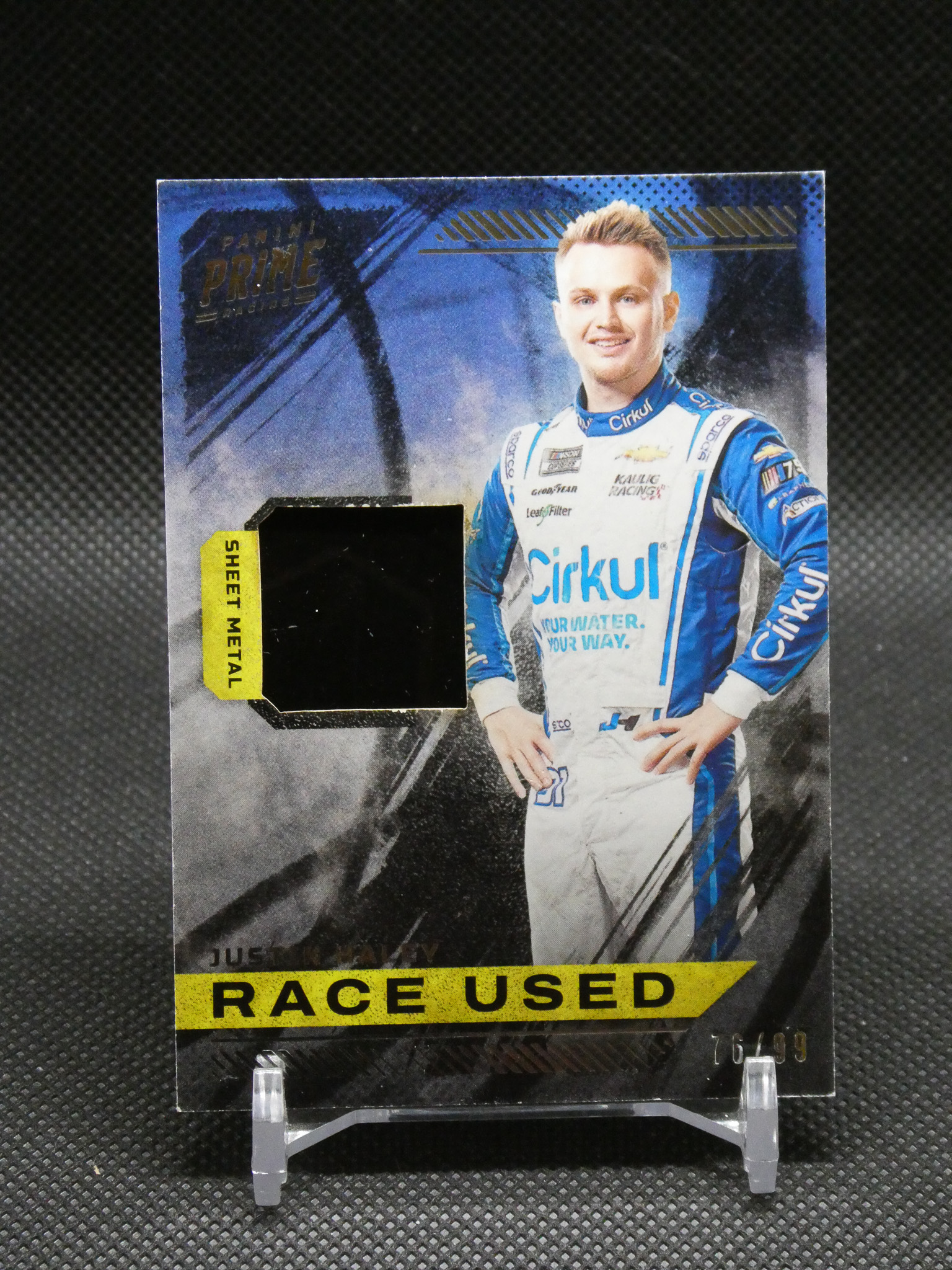 Promotional card showing a race car driver in a blue and white racing suit, with a yellow 'RACE USED' banner along the bottom.