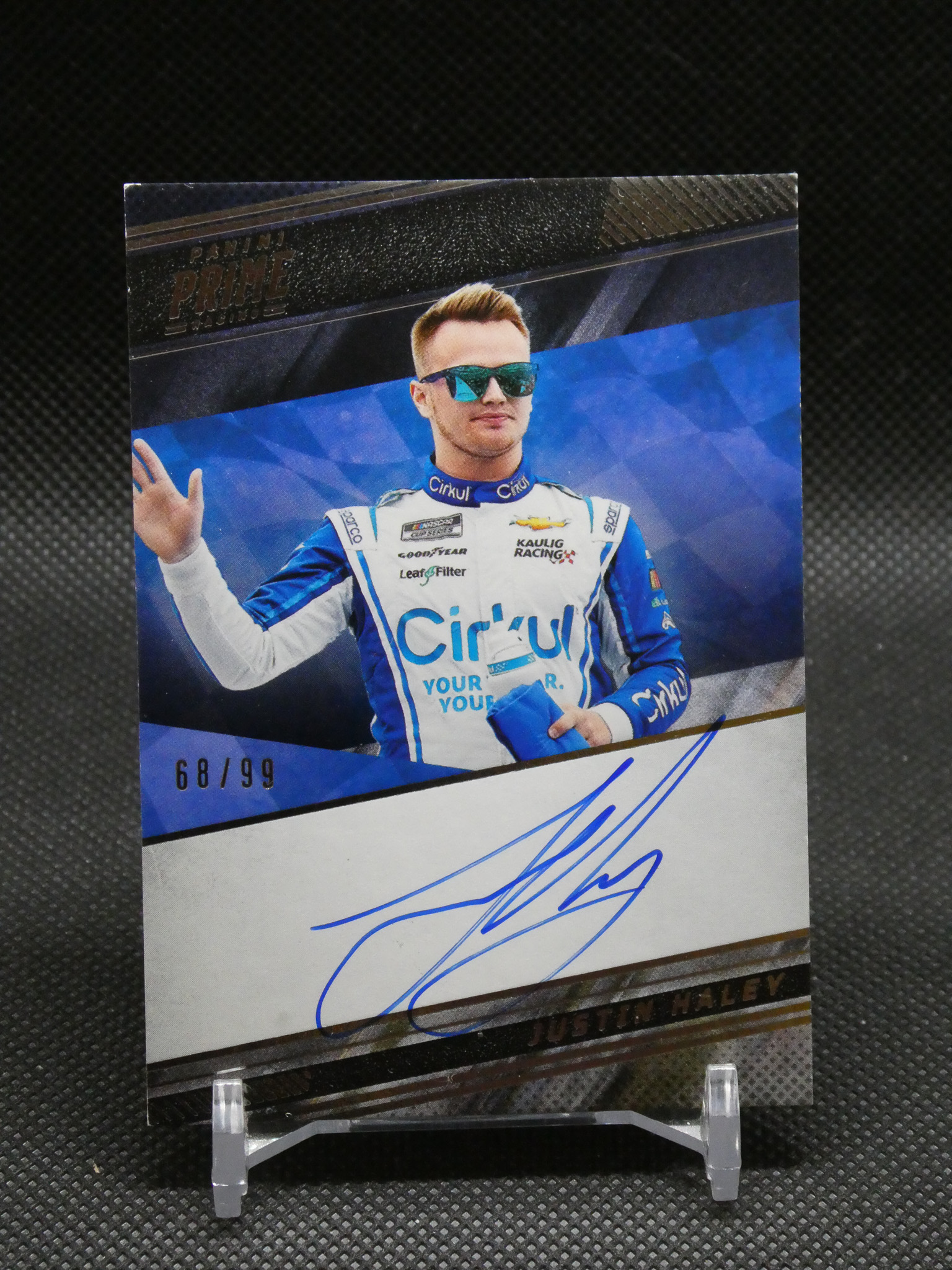 Portrait of a race car driver in a blue-and-white suit with mirrored sunglasses, waving from a signed poster on display.