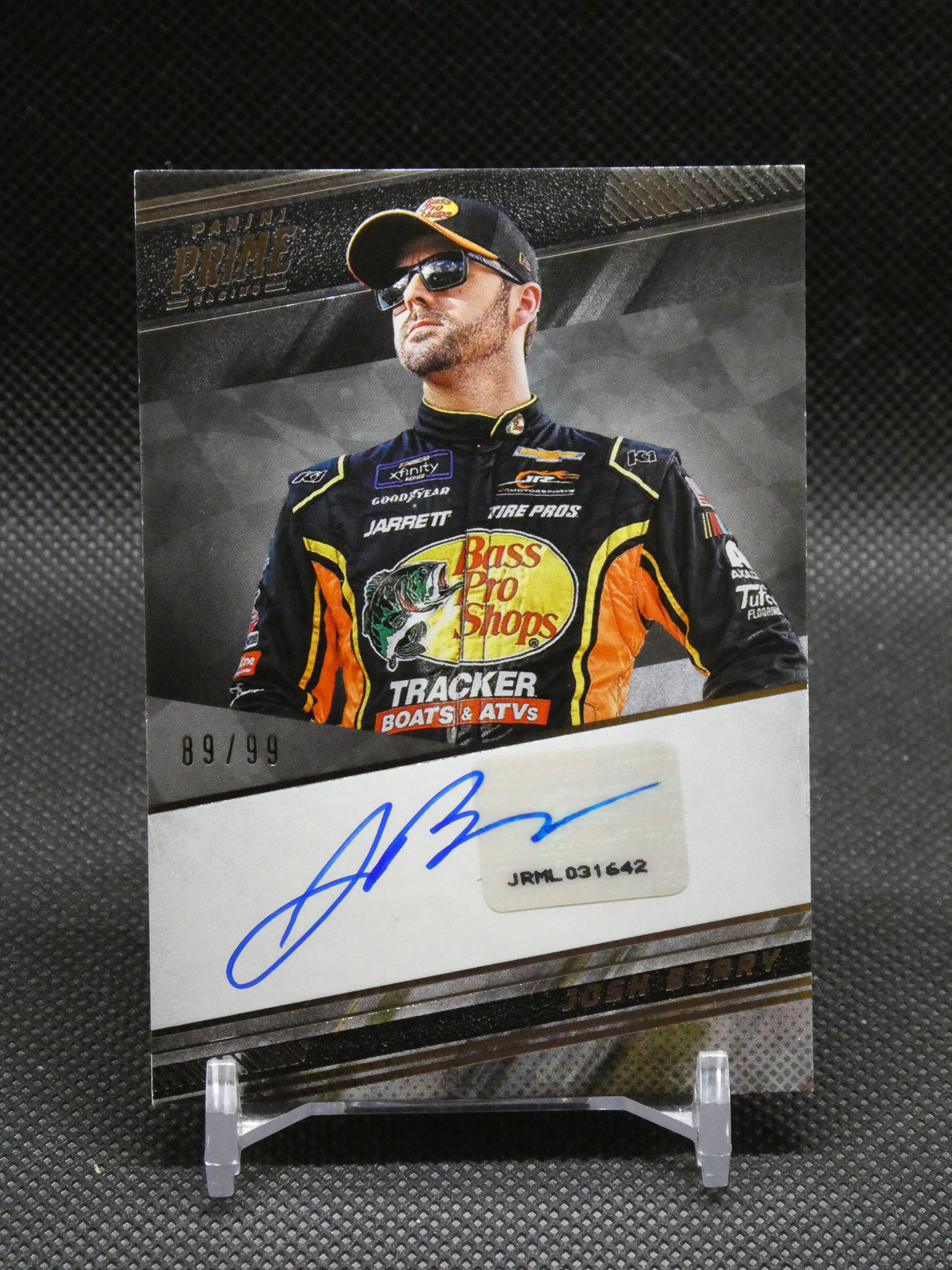 Autographed race poster of a driver in a black and yellow suit with Bass Pro Shops logo, wearing a cap and sunglasses, numbered 89/99 on a display stand.