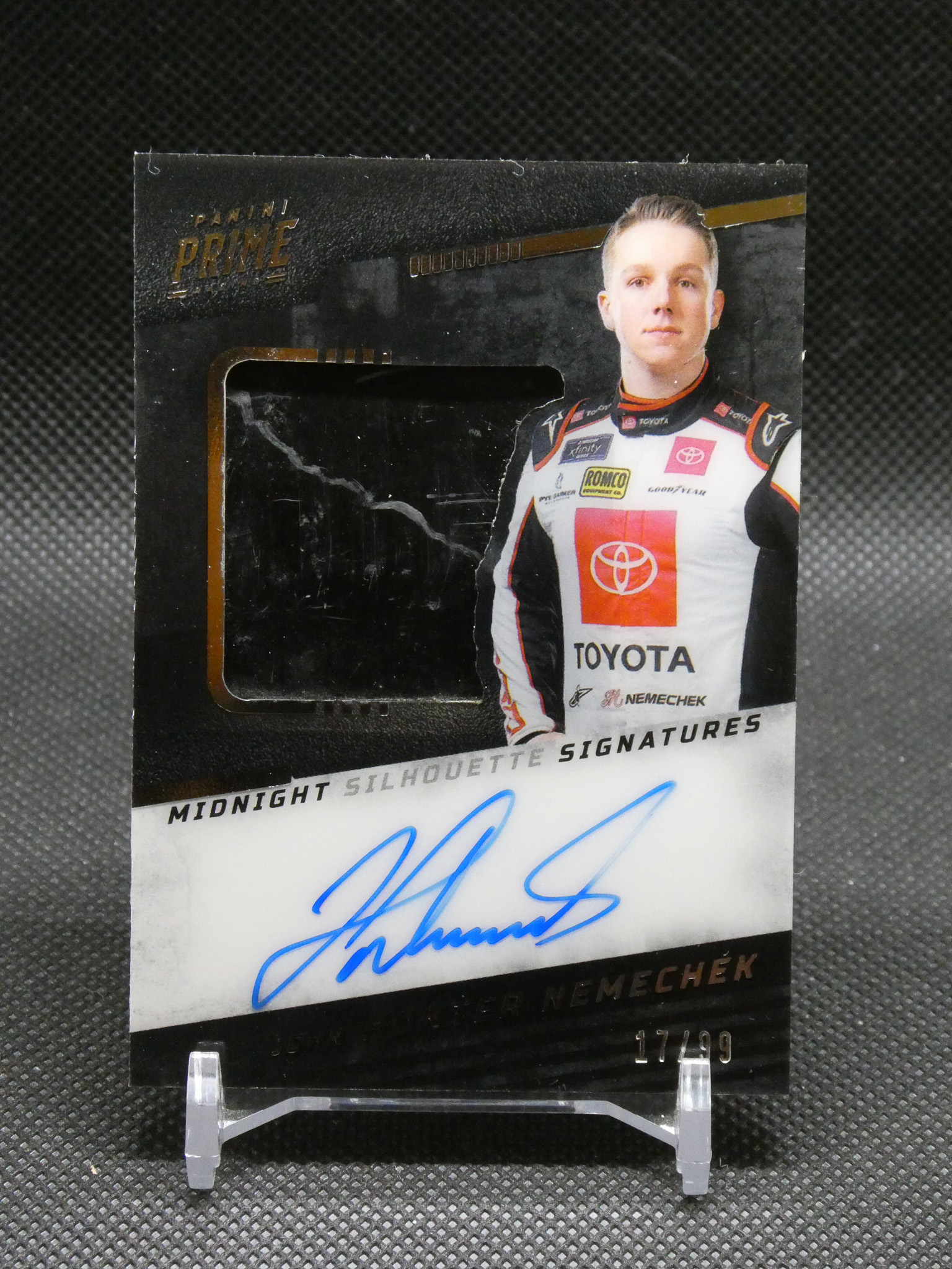 Autographed racing card featuring a driver in a white Toyota racing suit with team logos, mounted on a black backing and showing a blue signature at the bottom edge.