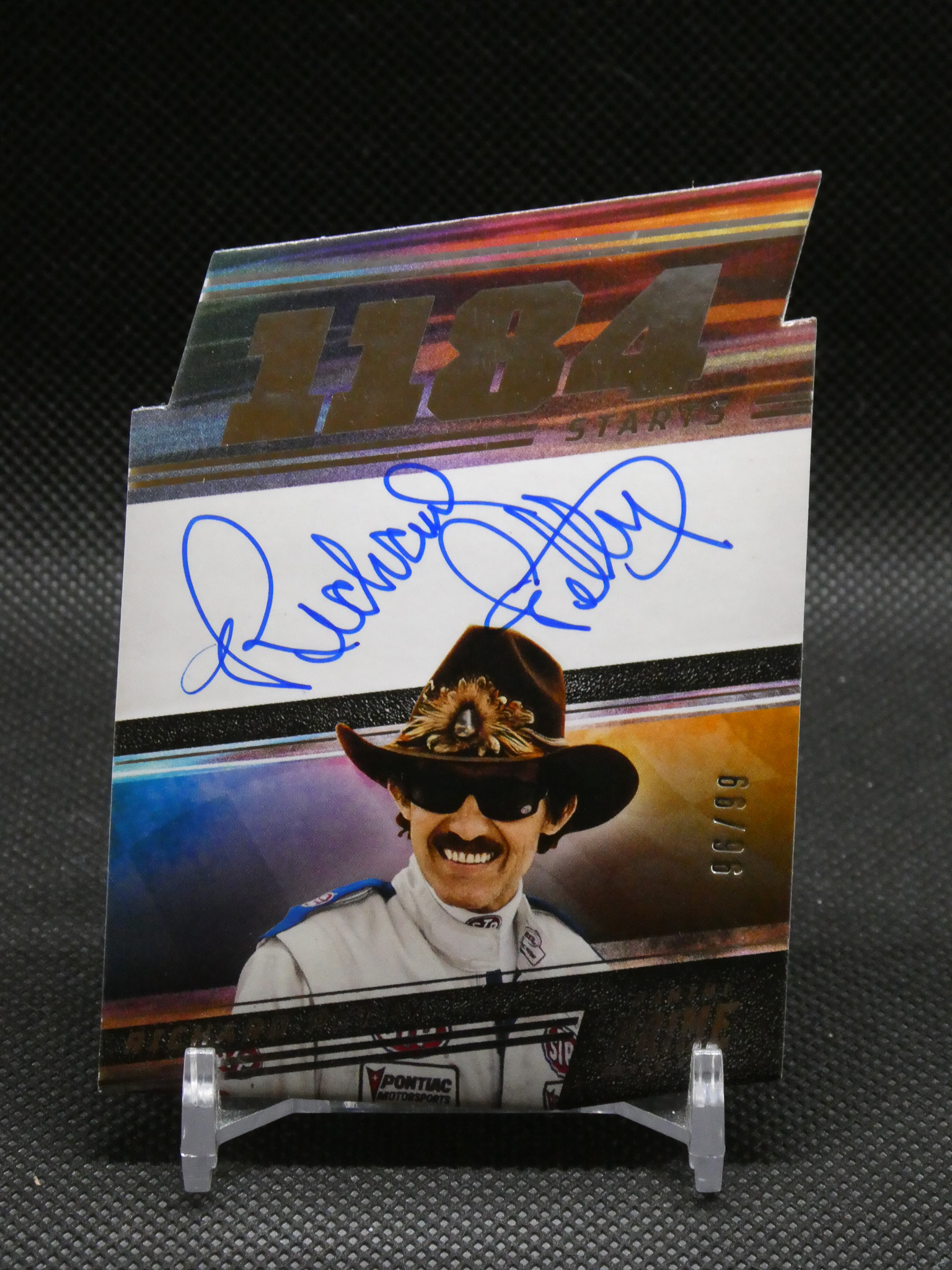 Autographed poster of a smiling race driver in a white suit wearing a cowboy hat and sunglasses, with a blue signature across the image, displayed on a clear stand.