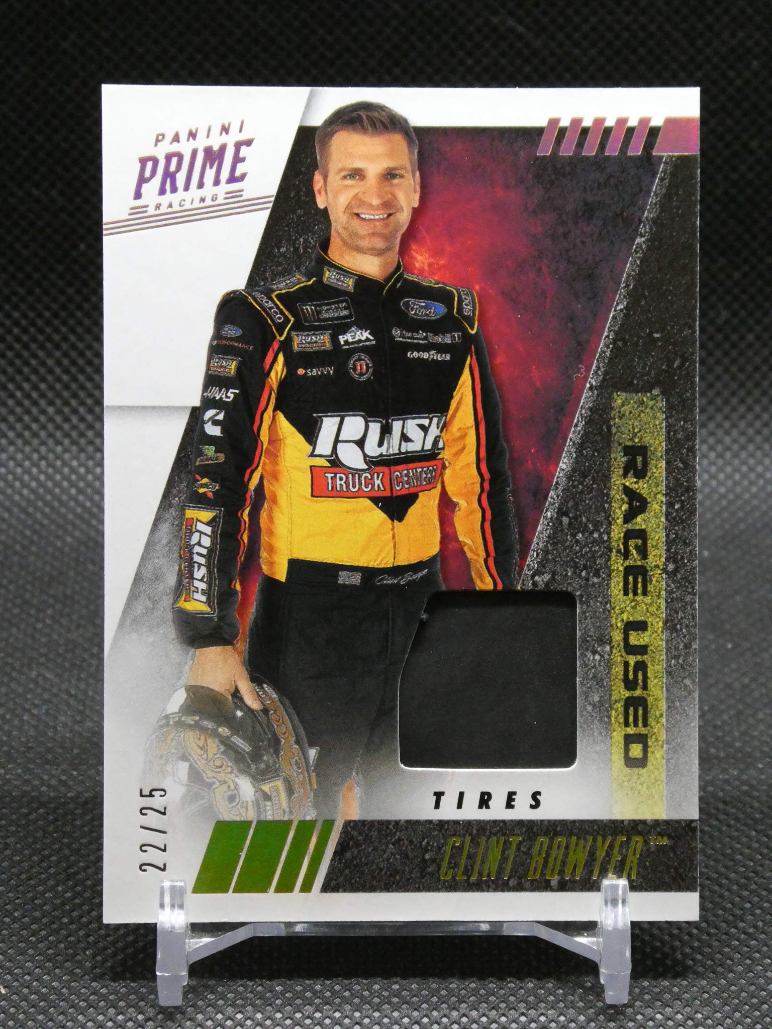 Trading card of NASCAR driver Clint Bowyer in a yellow-black racing suit, Panini Prime Racing logo top left, bold red/black background, smiling.