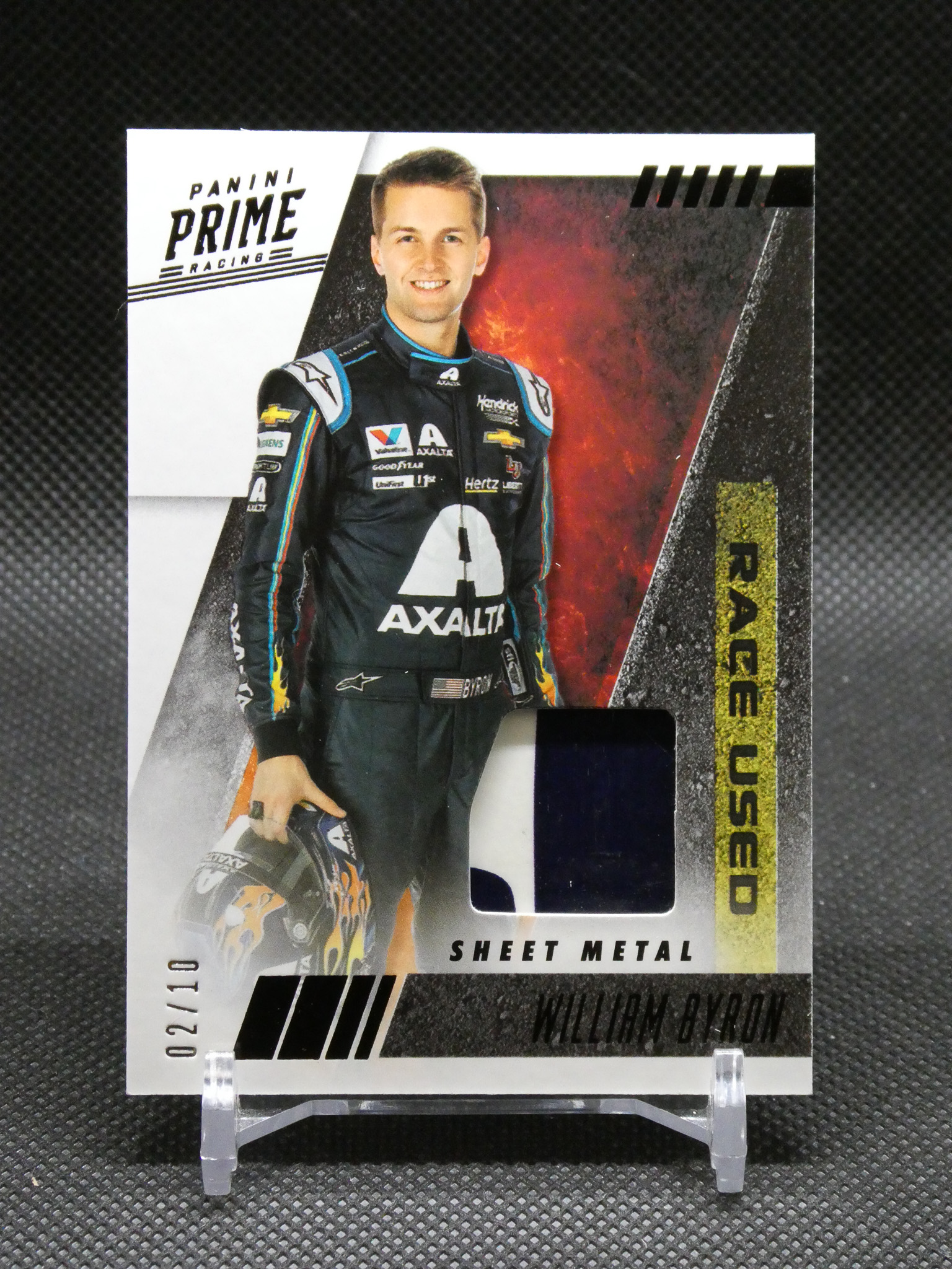 Panini Prime Racing trading card featuring driver William Byron in a black Axalta suit, holding his helmet on a display stand.