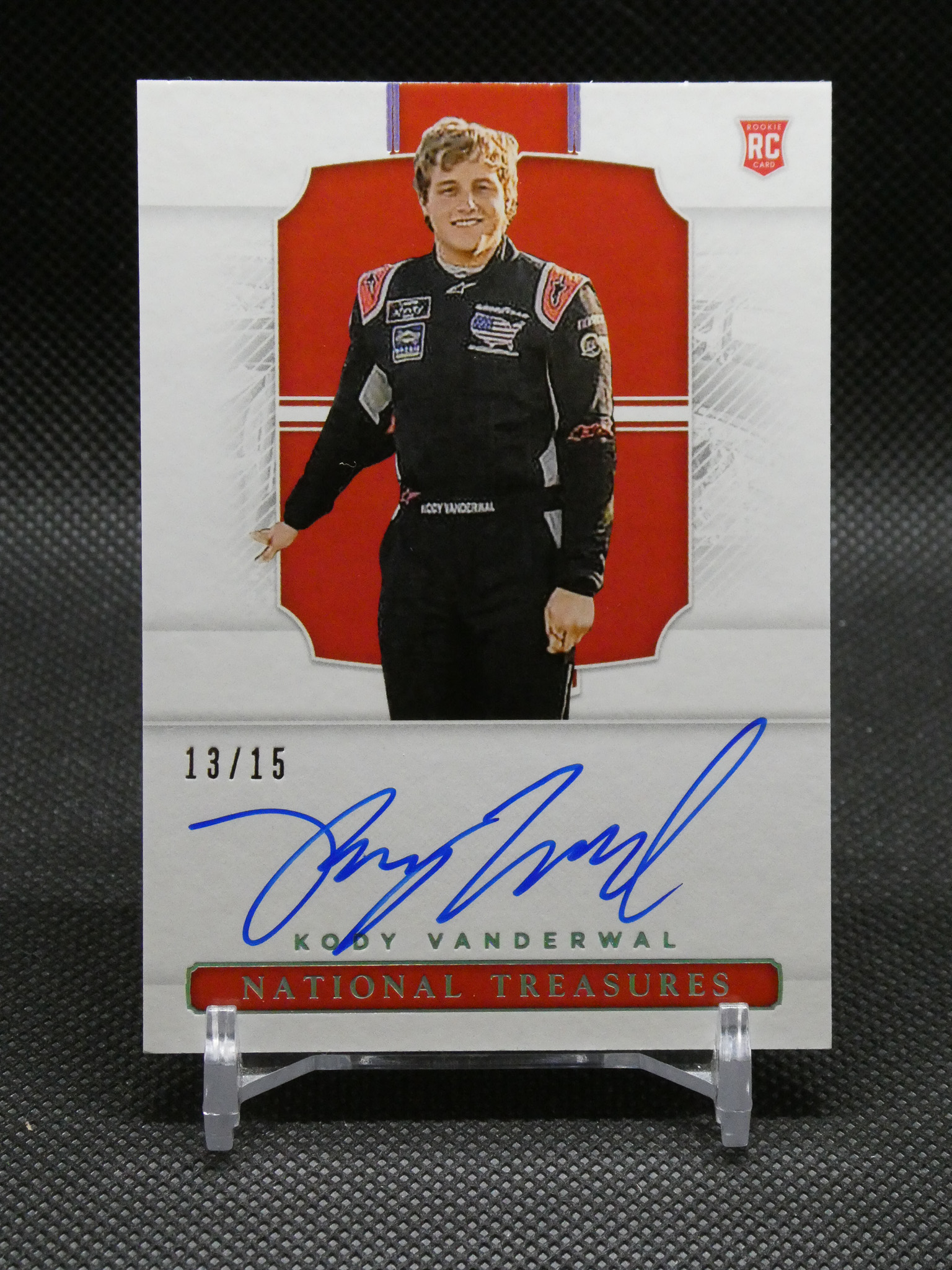 Signed trading card featuring a race car driver in a black racing suit, standing in front of a red backdrop; edition 13/15, National Treasures branding visible at the bottom.