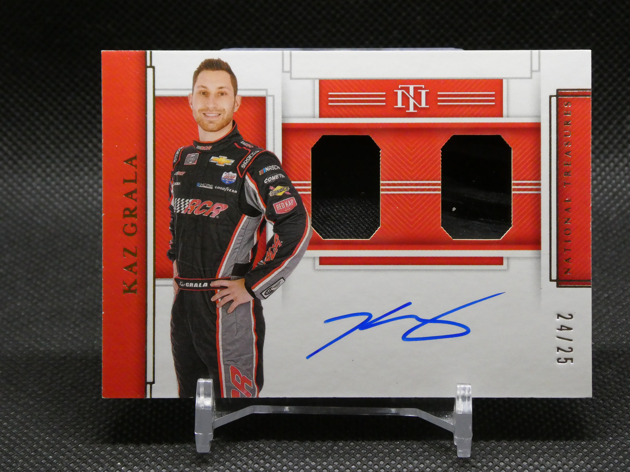 Portrait of a male race car driver in a black suit with sponsor logos, standing beside a red and white display backdrop.