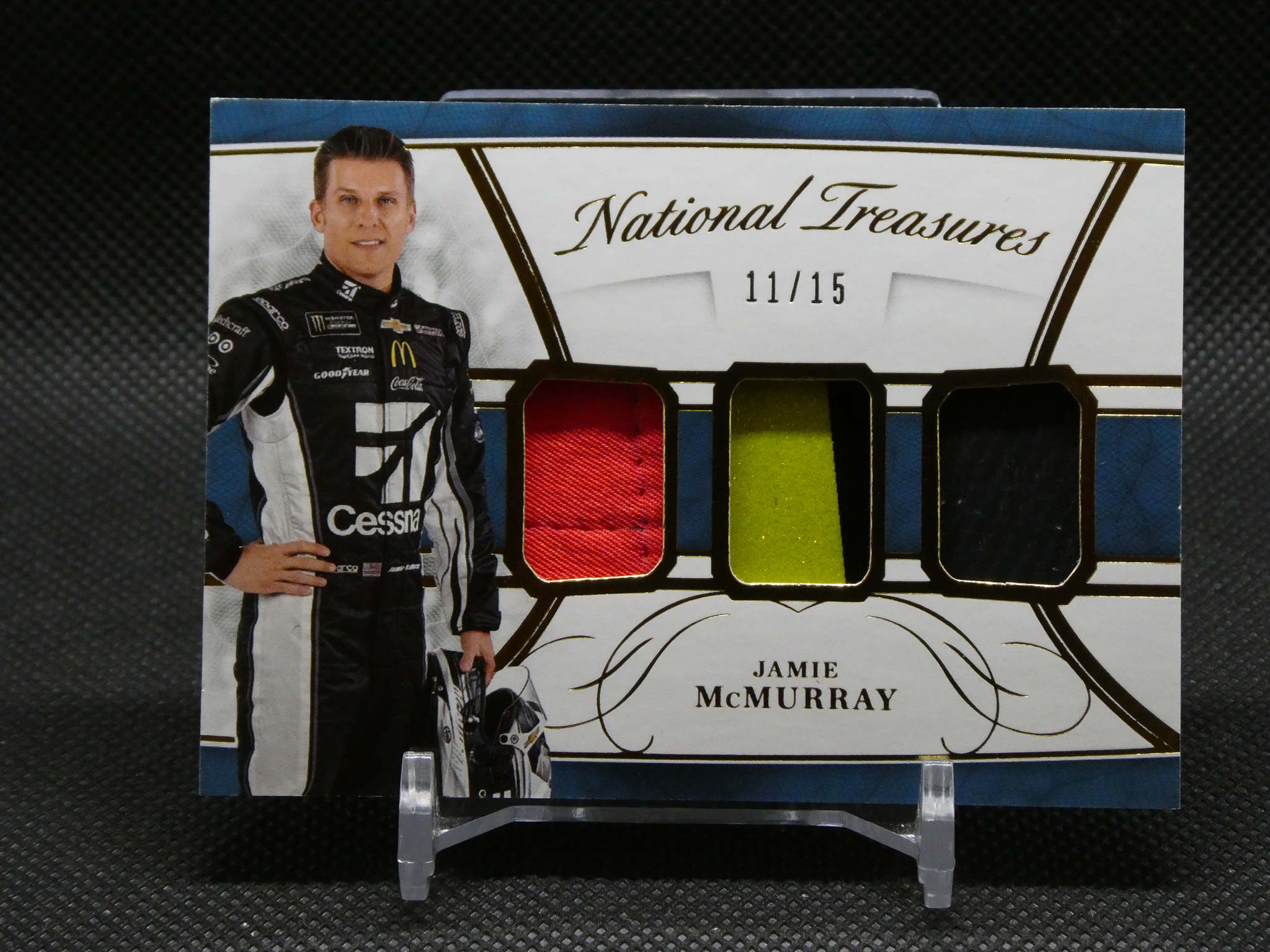 Portrait of NASCAR driver Jamie McMurray in a racing suit standing beside a display labeled 'National Treasures' with color swatches (red, yellow, green).