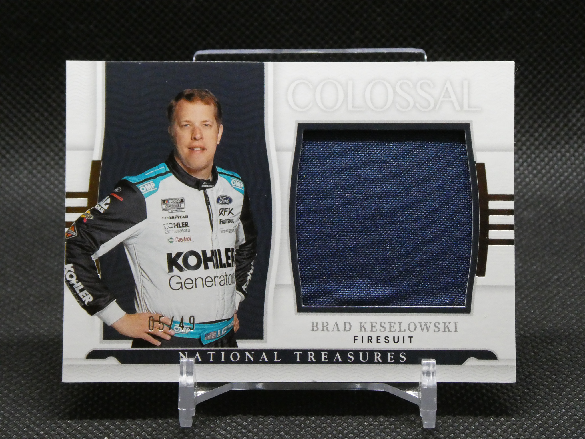 Brad Keselowski in a white racing suit stands beside a dark blue fabric swatch on a display card labeled Colossal and National Treasures
