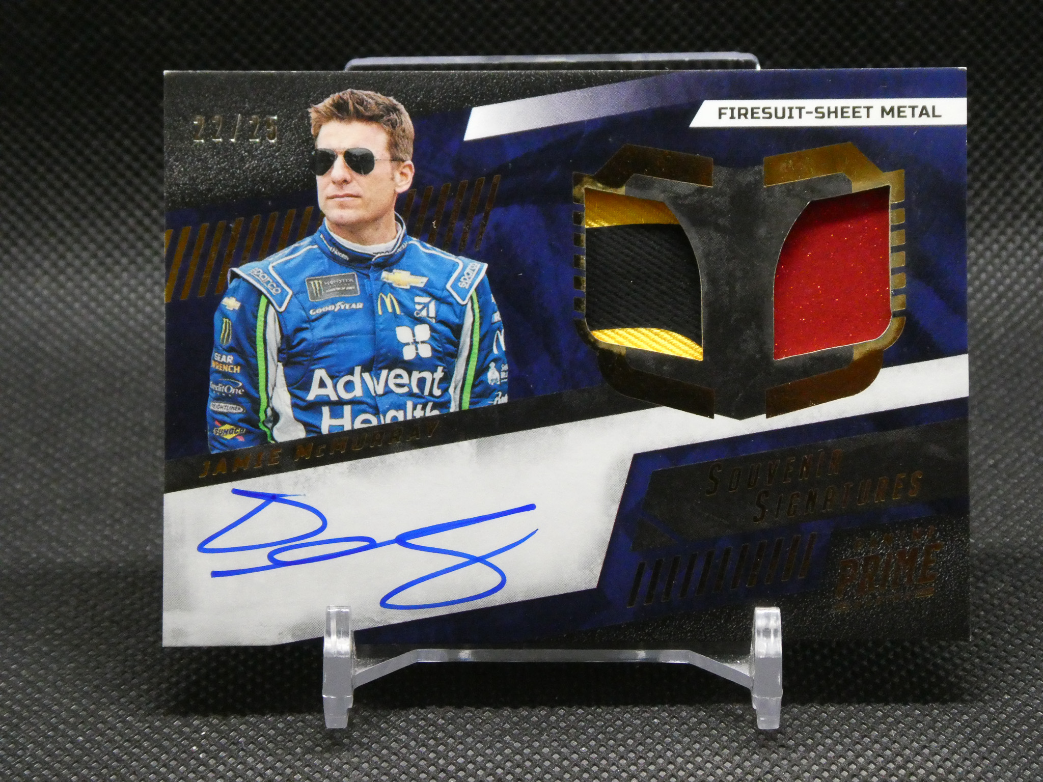 Trading card packaging featuring a NASCAR driver in a blue racing suit with sunglasses, next to a red gemstone graphic and a blue autograph at the bottom.