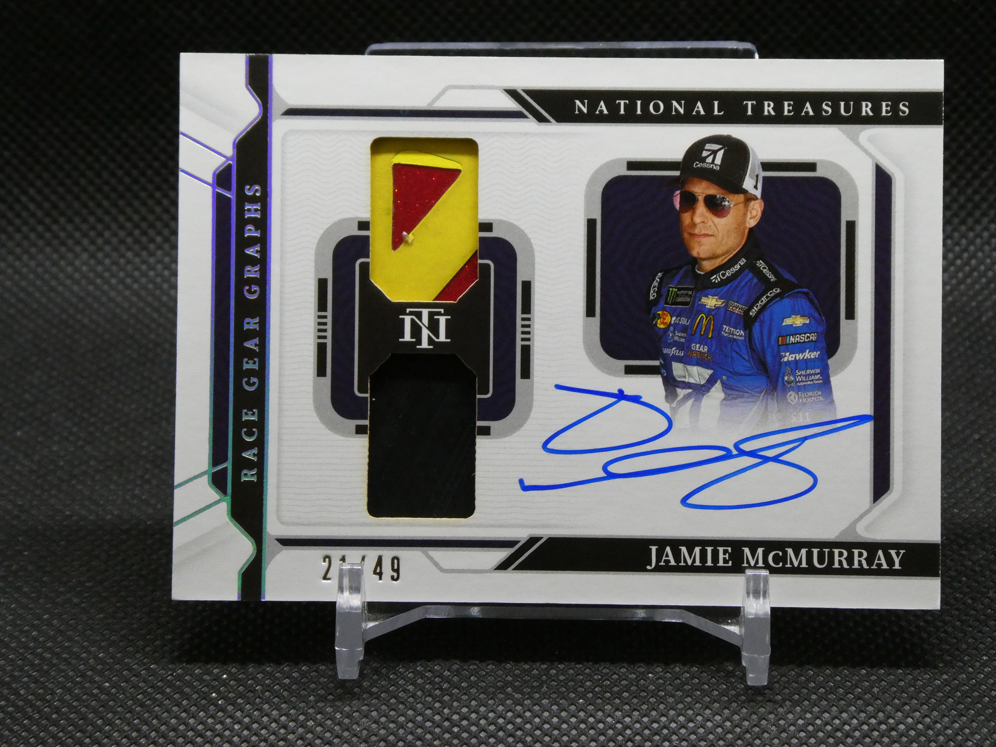 Close-up of a Jamie McMurray NASCAR trading card with a yellow-red fabric patch inset, autograph in blue ink, and 'National Treasures' branding visible in the background.