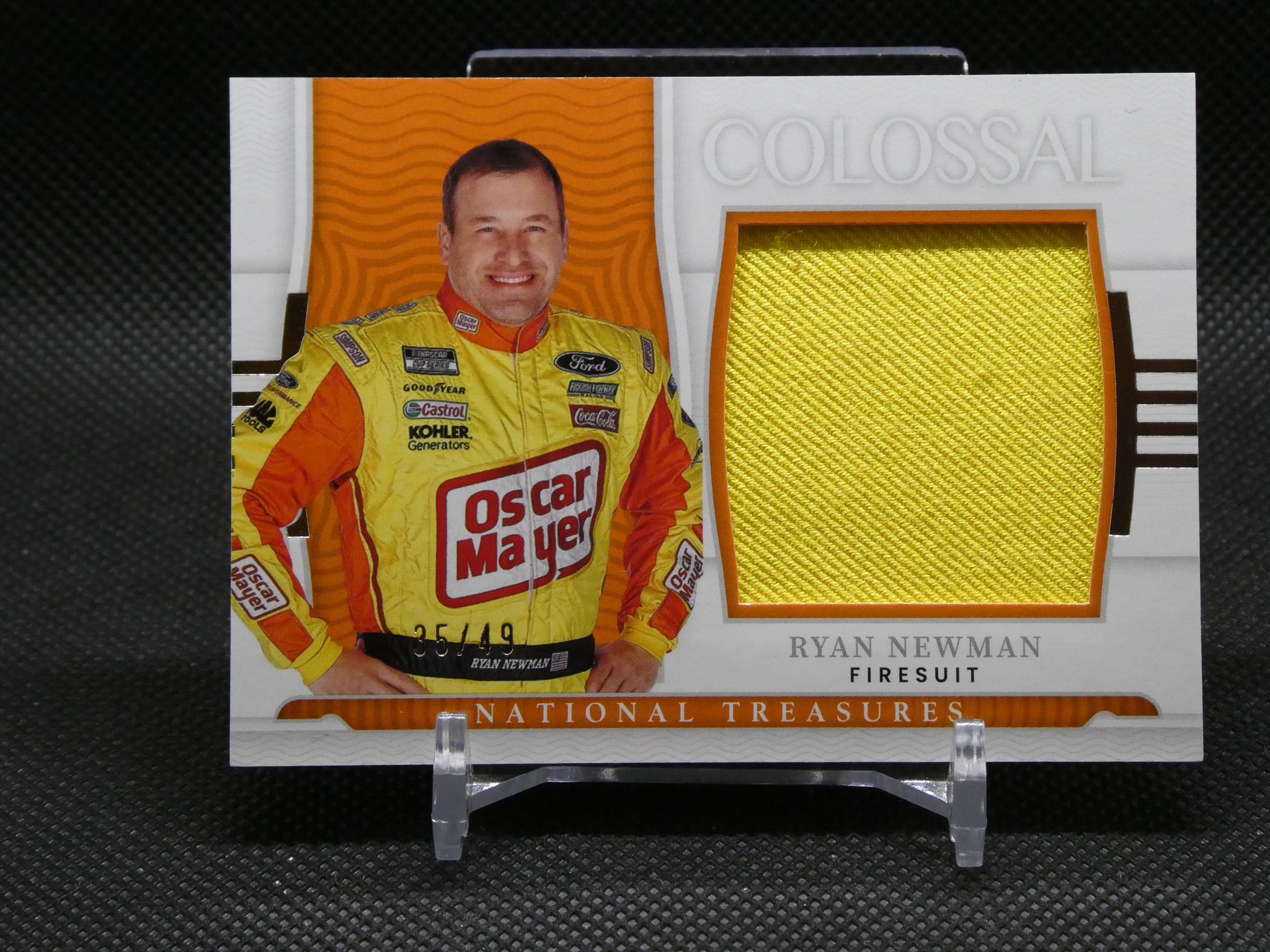 Portrait of NASCAR driver Ryan Newman in a yellow sponsor-laden firesuit, standing beside a textured gold fabric swatch on a display card.