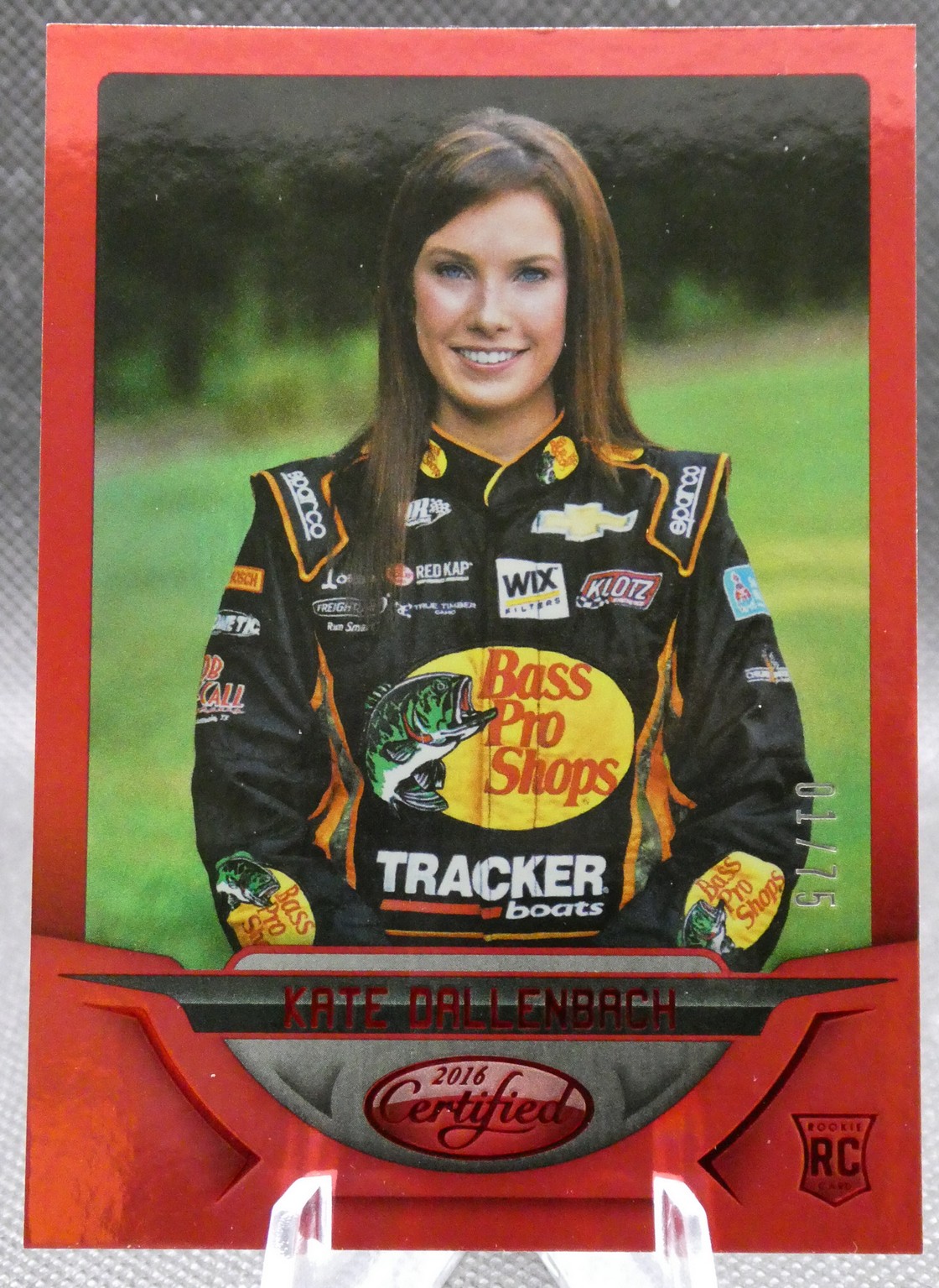Portrait of race car driver Kate Dallenbach in a black and orange Bass Pro Shops racing suit, smiling outdoors with a red-bordered card frame.