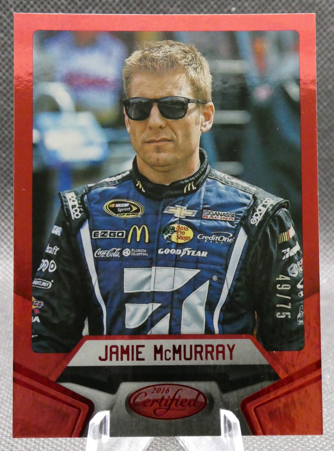Jamie McMurray in a blue racing suit with sunglasses, standing for a trading card on a red border.