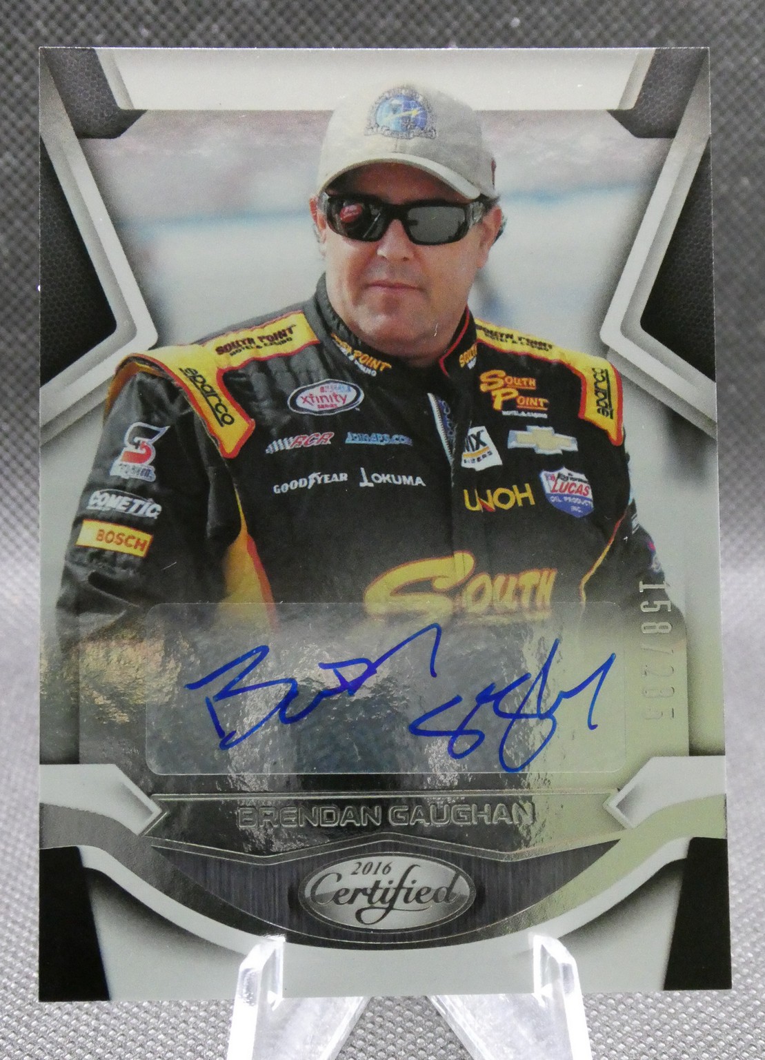 Autographed 2016 Certified trading card featuring NASCAR driver Brendan Gaughan in a black and yellow sponsor-laden racing suit, wearing sunglasses.