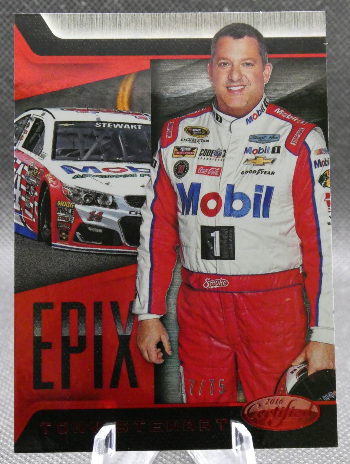 Portrait of NASCAR driver Tony Stewart in a white Mobil 1 racing suit, standing beside a red–white race car with 'Stewart' on the windshield backdrop.