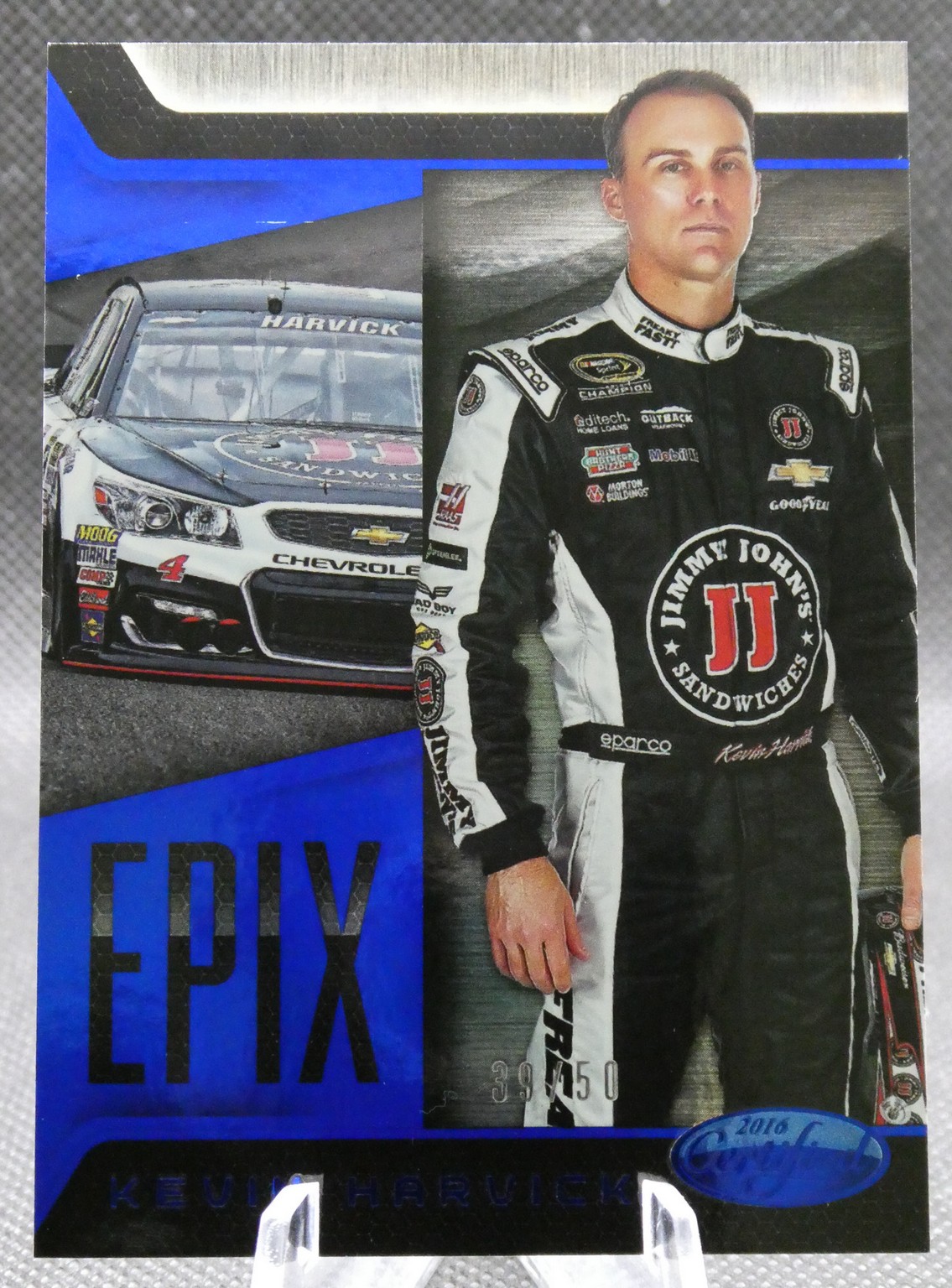 Race car driver in a black-and-white sponsor-branded racing suit standing beside a Chevrolet race car on track surface.
