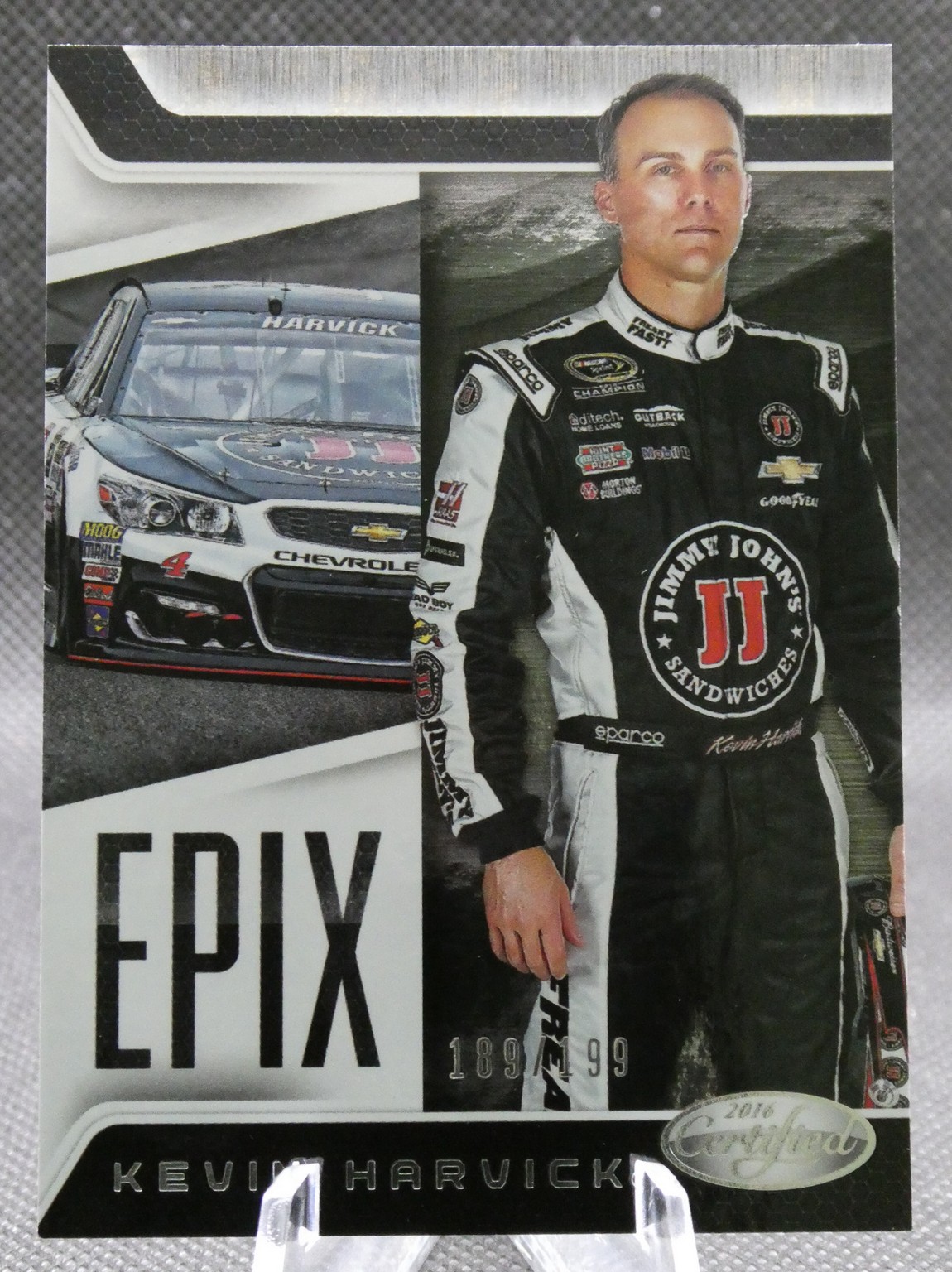 Portrait of Kevin Harvick in a black-and-white racing suit beside a Chevrolet race car with sponsor logos, on a poster backdrop.