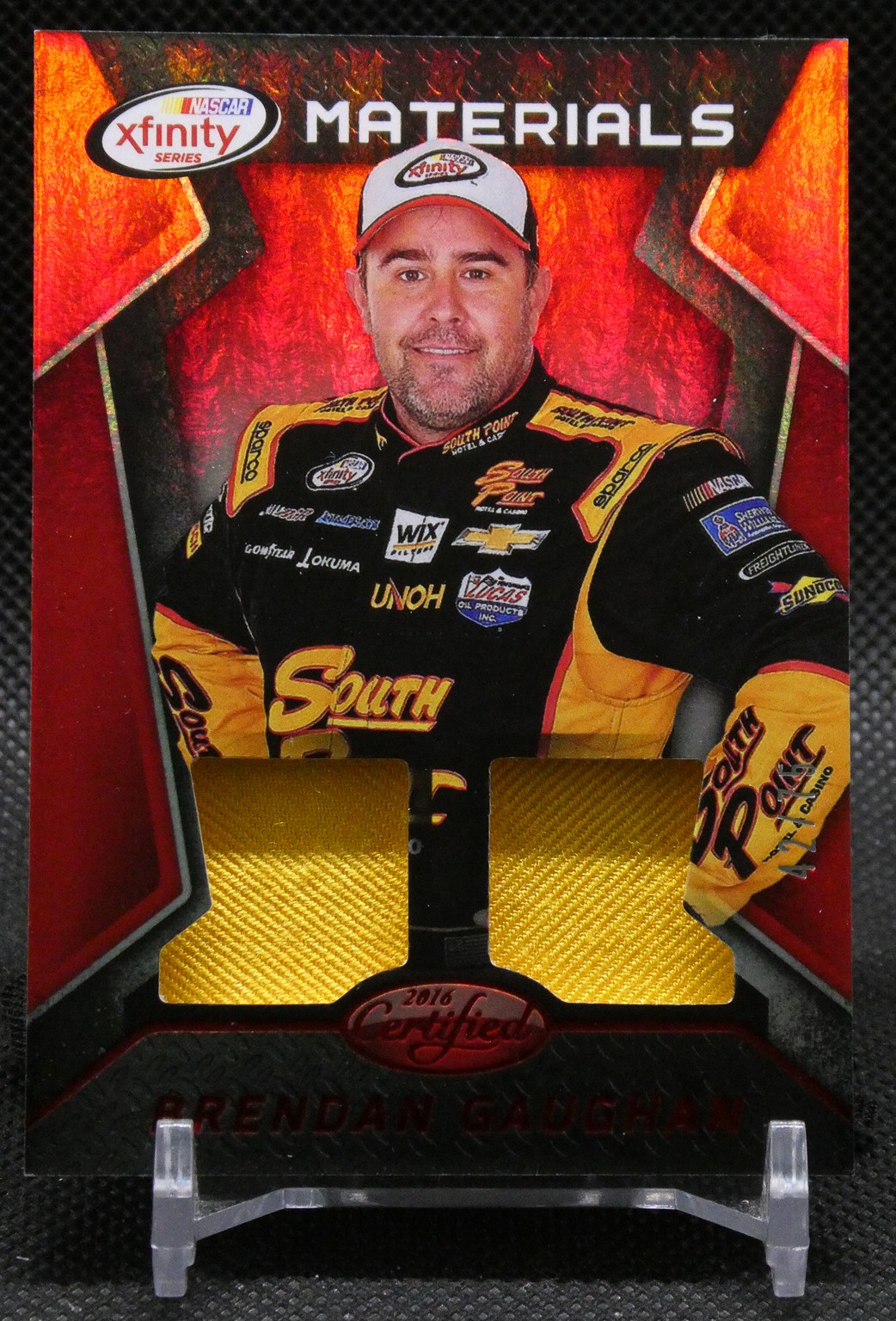 NASCAR Xfinity Materials trading card showing a driver in a black-yellow suit with two gold fabric swatches and sponsor logos, 'Materials' header at top.