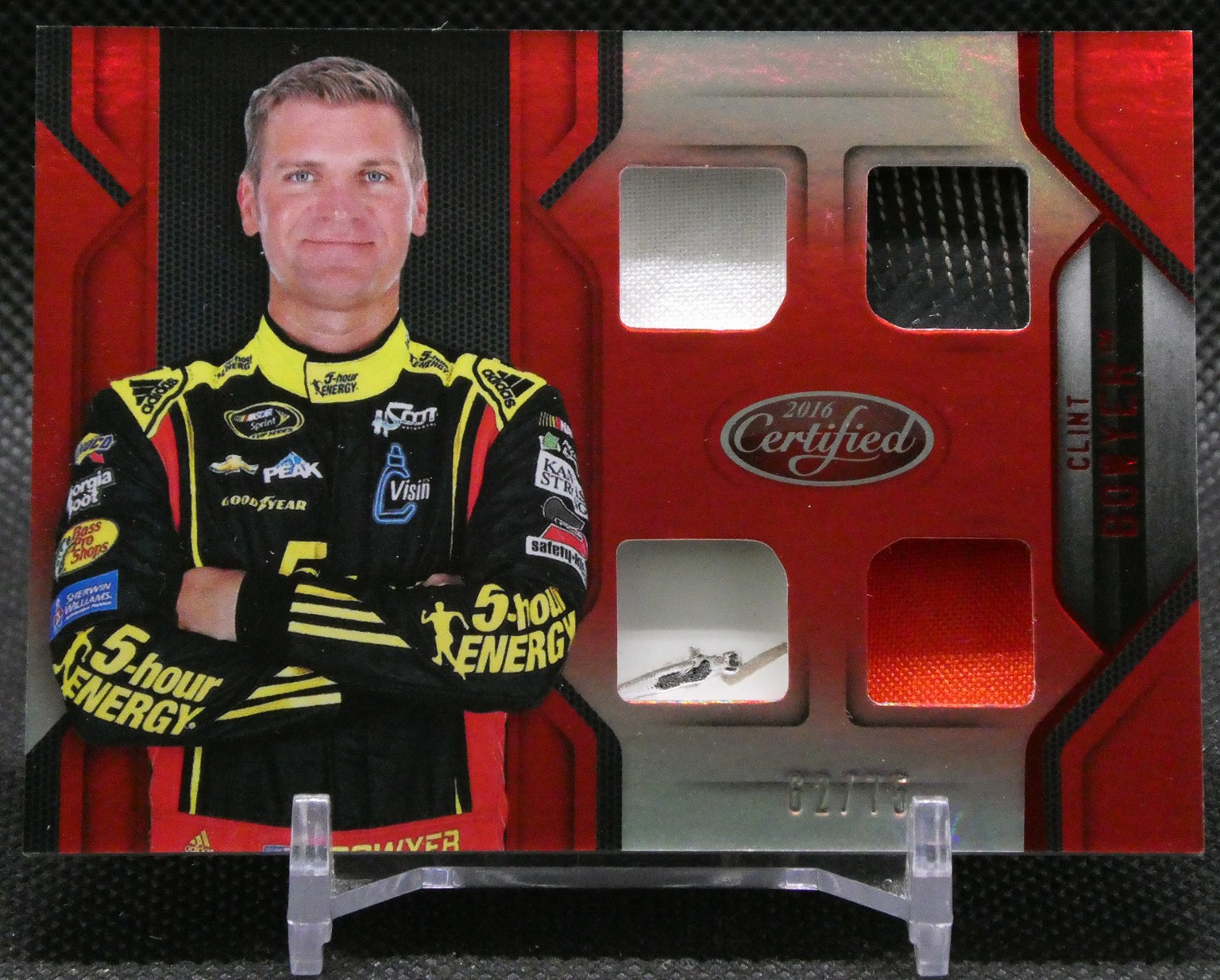 Trading card of a race car driver in a yellow-and-black sponsor suit, arms crossed, with a red panel background and fabric swatches nearby.