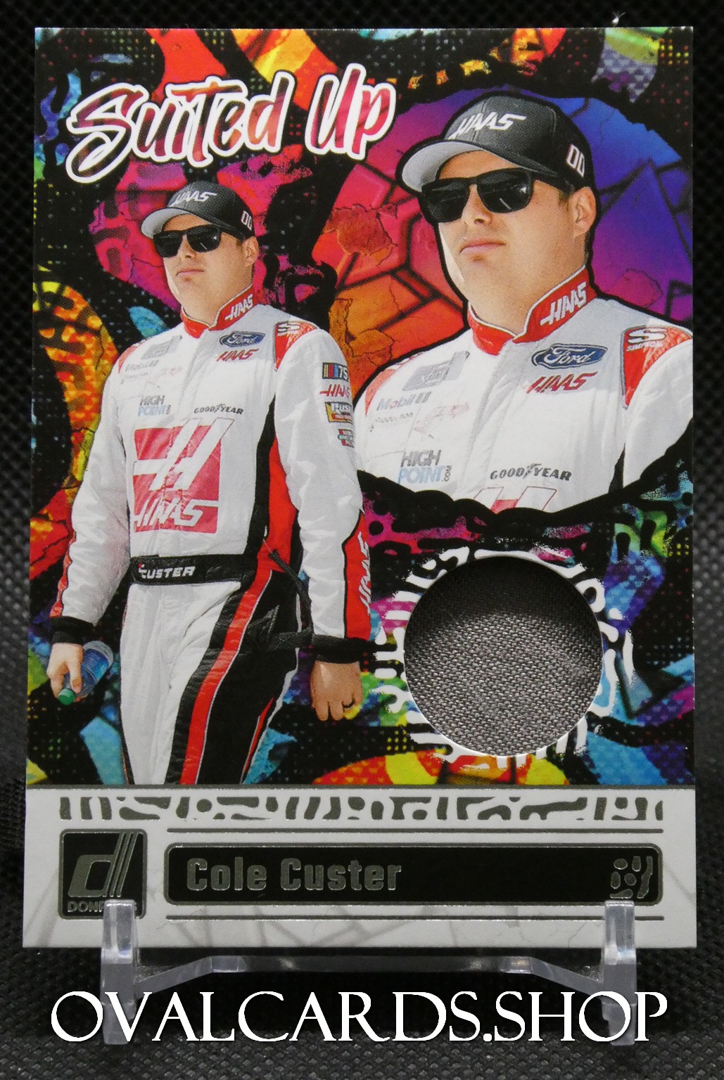 Trading card featuring NASCAR driver Cole Custer in a white and red racing suit, wearing sunglasses and a cap, with a colorful graffiti background and a circular fabric relic window near the bottom.