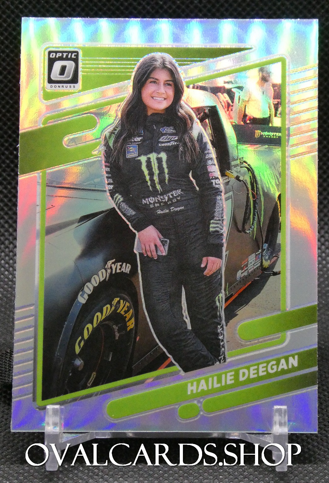 Trading card-style image of a female race car driver in a black Monster Energy suit standing beside a race car with Goodyear tires, on a holographic card backdrop.