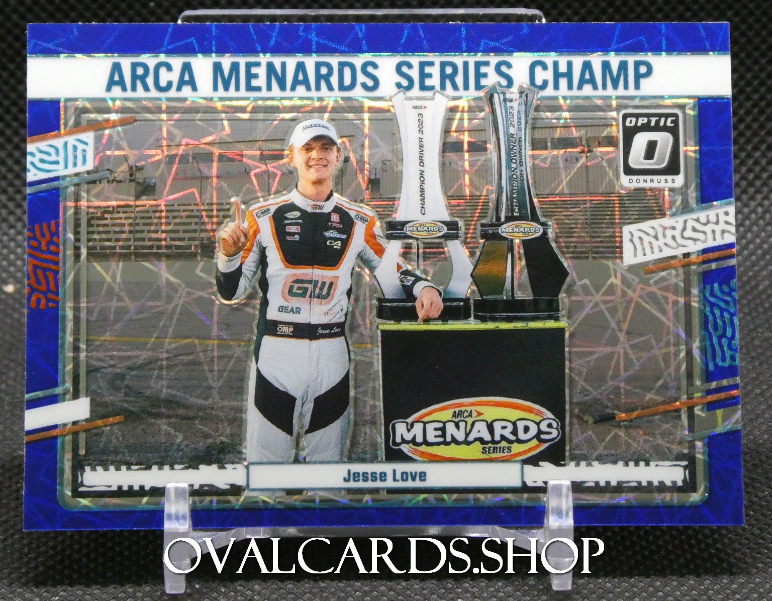 Driver in racing suit posing with ARCA Menards Series champion trophies, banner above reads ARCA MENARDS SERIES CHAMP.