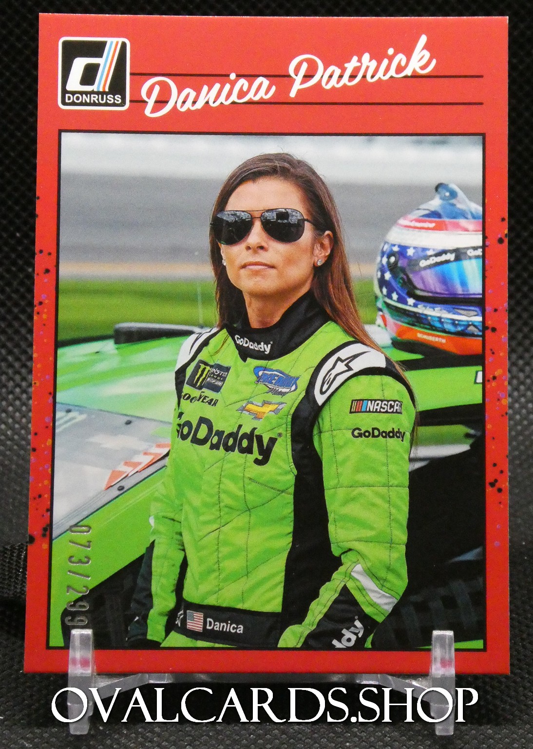 Portrait of Danica Patrick in a bright green GoDaddy racing suit, standing next to a racing car with a helmet in the background.