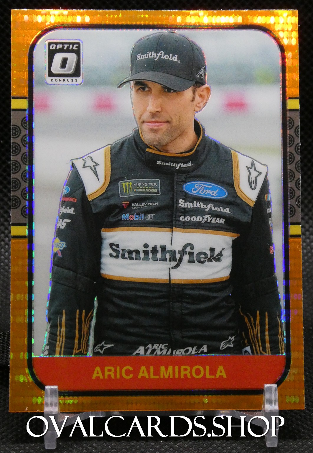 Trading card image of NASCAR driver Aric Almirola in a black Smithfield Ford racing suit and cap, orange border visible, with his name at the bottom.