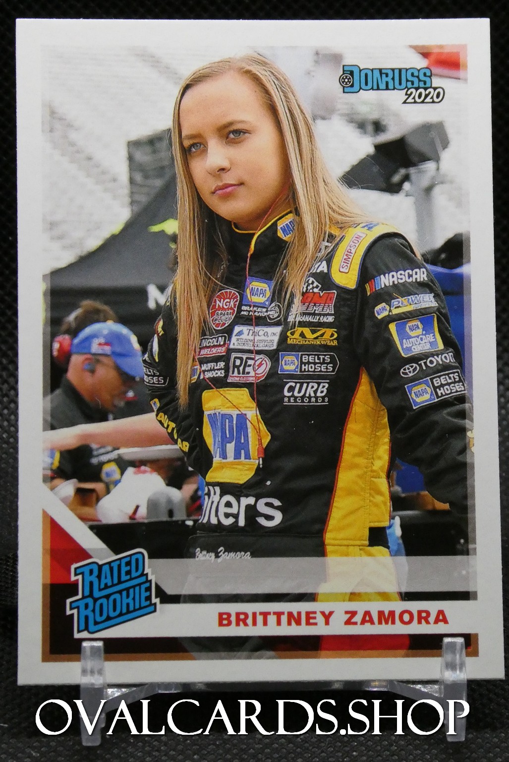 Trading card-style photo of Brittney Zamora in a black and yellow NASCAR jumpsuit with sponsor patches, standing at a racing event; bottom shows her name and a 'Rated Rookie' badge.