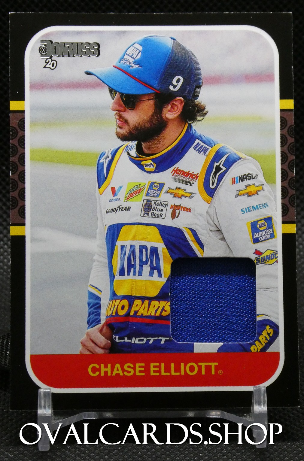 Chase Elliott trading card displaying a portrait of the NASCAR driver in a blue-yellow racing suit with a blue fabric swatch embedded on the card and sponsor logos visible behind him, including Chevrolet and Hendrick Motorsports.