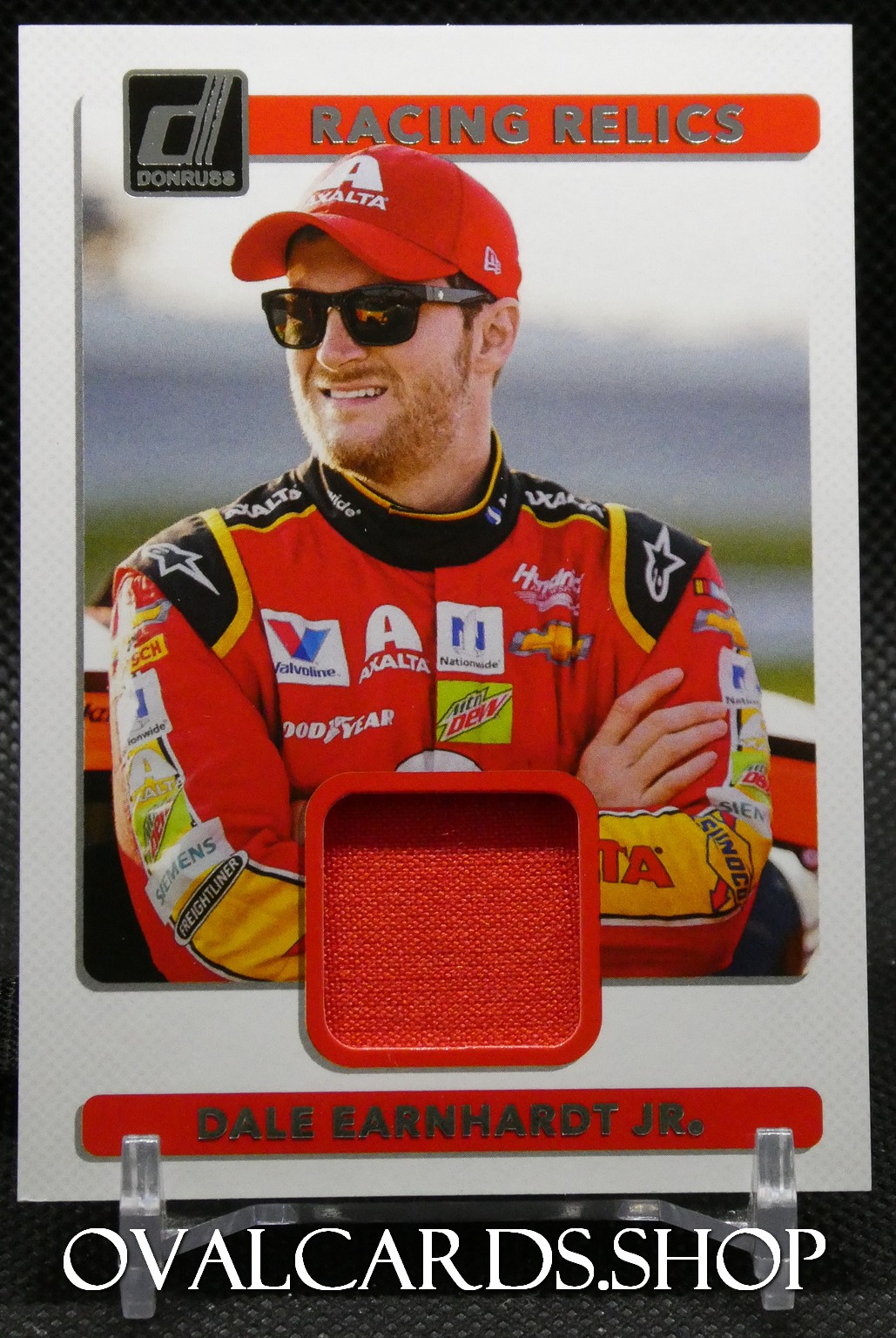 Trading card featuring NASCAR driver Dale Earnhardt Jr. in a red suit with a fabric relic inset and 'Racing Relics' header.