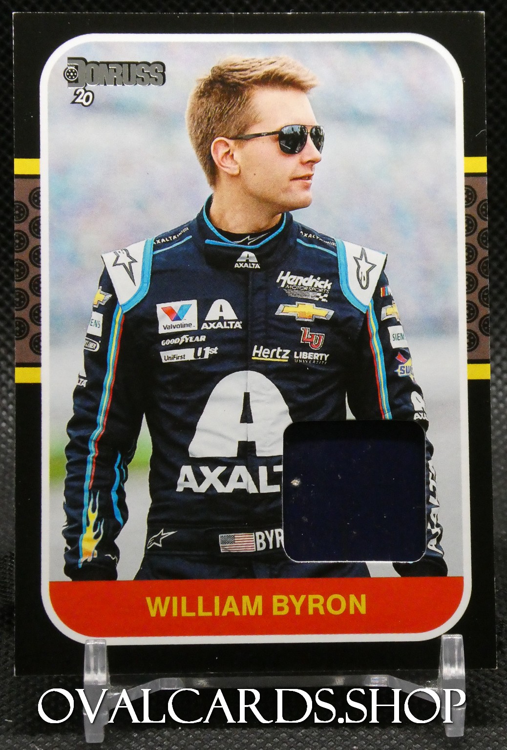 Portrait of William Byron in a dark sponsor-laden racing suit, wearing sunglasses, standing outdoors; includes Axalta logos and the name 'William Byron' on the card.